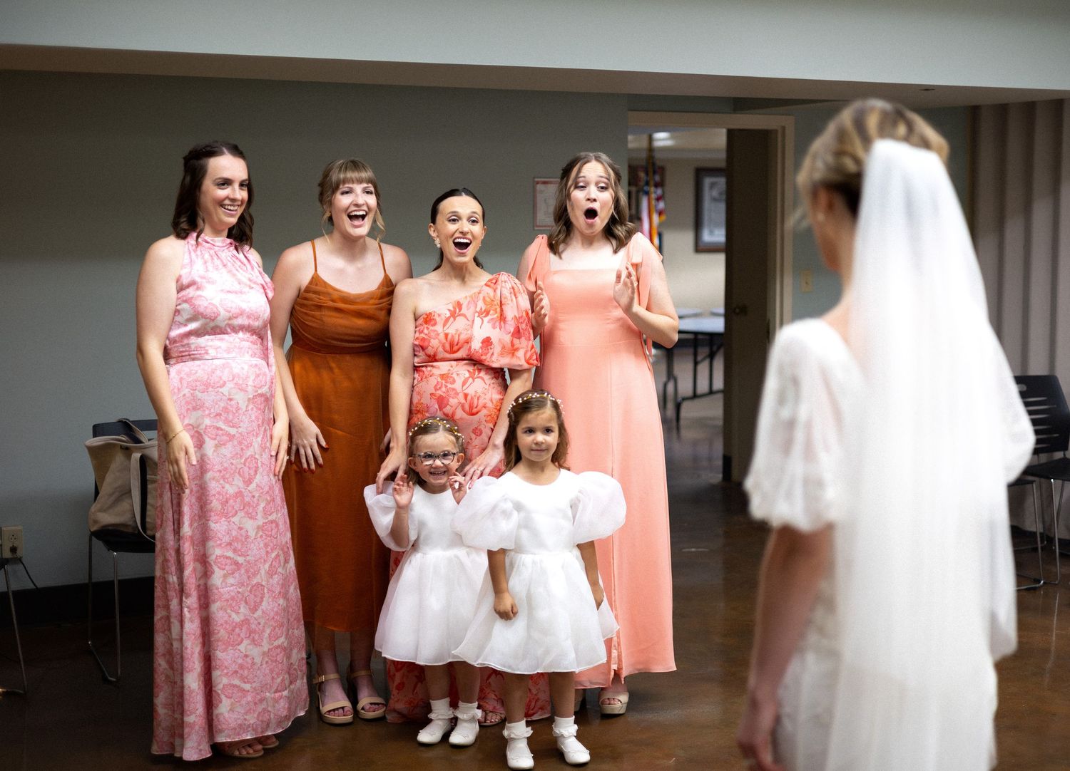 Wedding party members in coral dresses and white gowns pose together in an indoor setting.