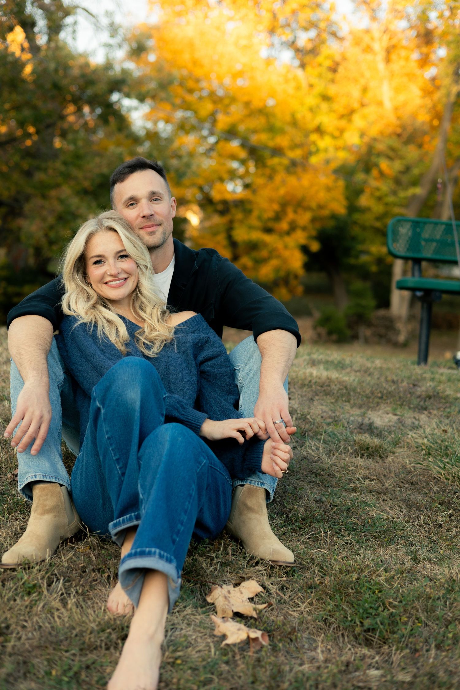 A couple in denim sits together on a grassy hill with golden autumn foliage in the background.