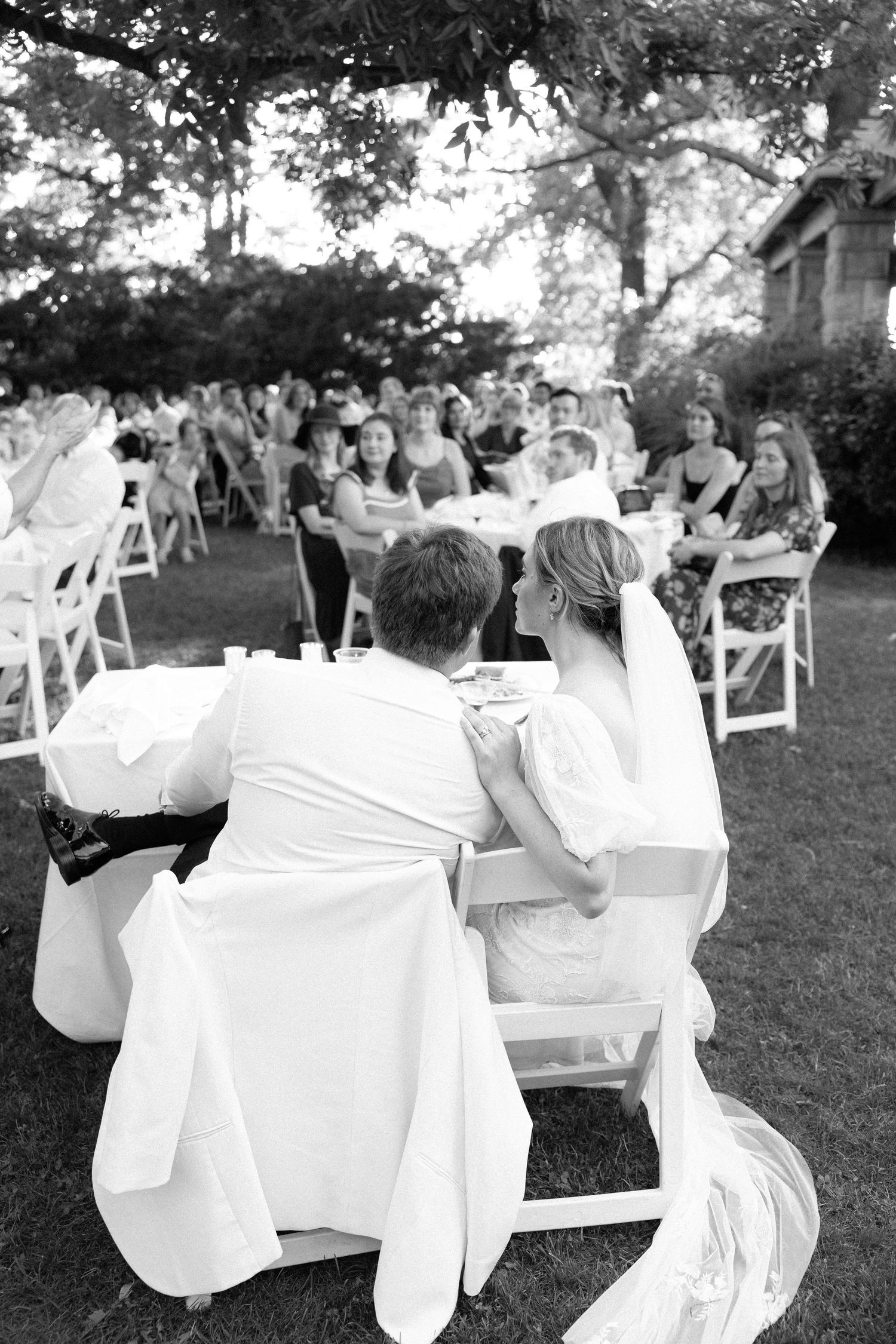 A wedding reception in a garden setting with white chairs and tables under trees.