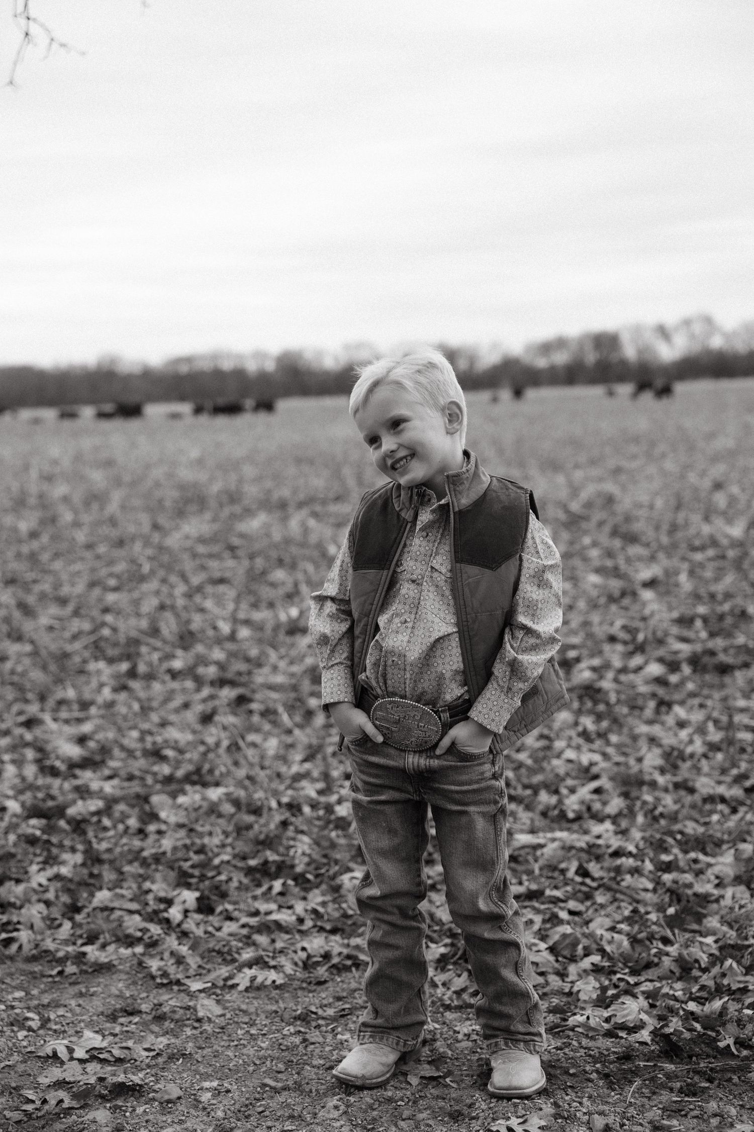 A young child in a vest and jeans stands in a rural field on an overcast day.