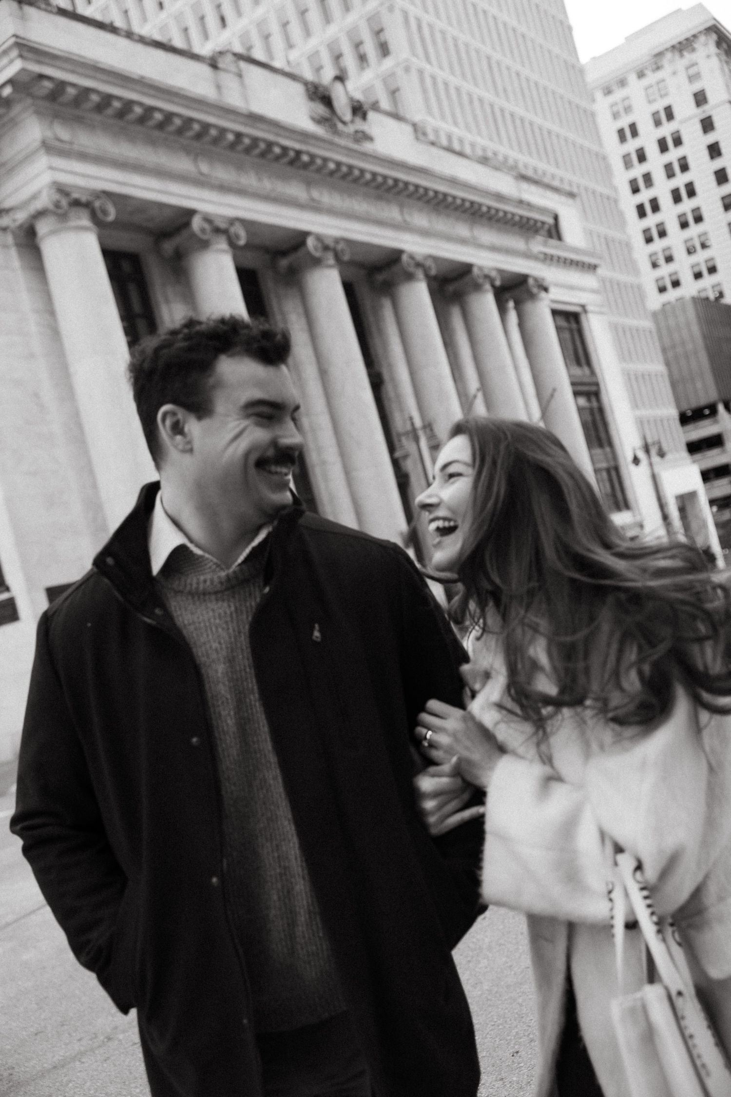 A romantic couple laughs and embraces while standing in front of a classical building with columns.
