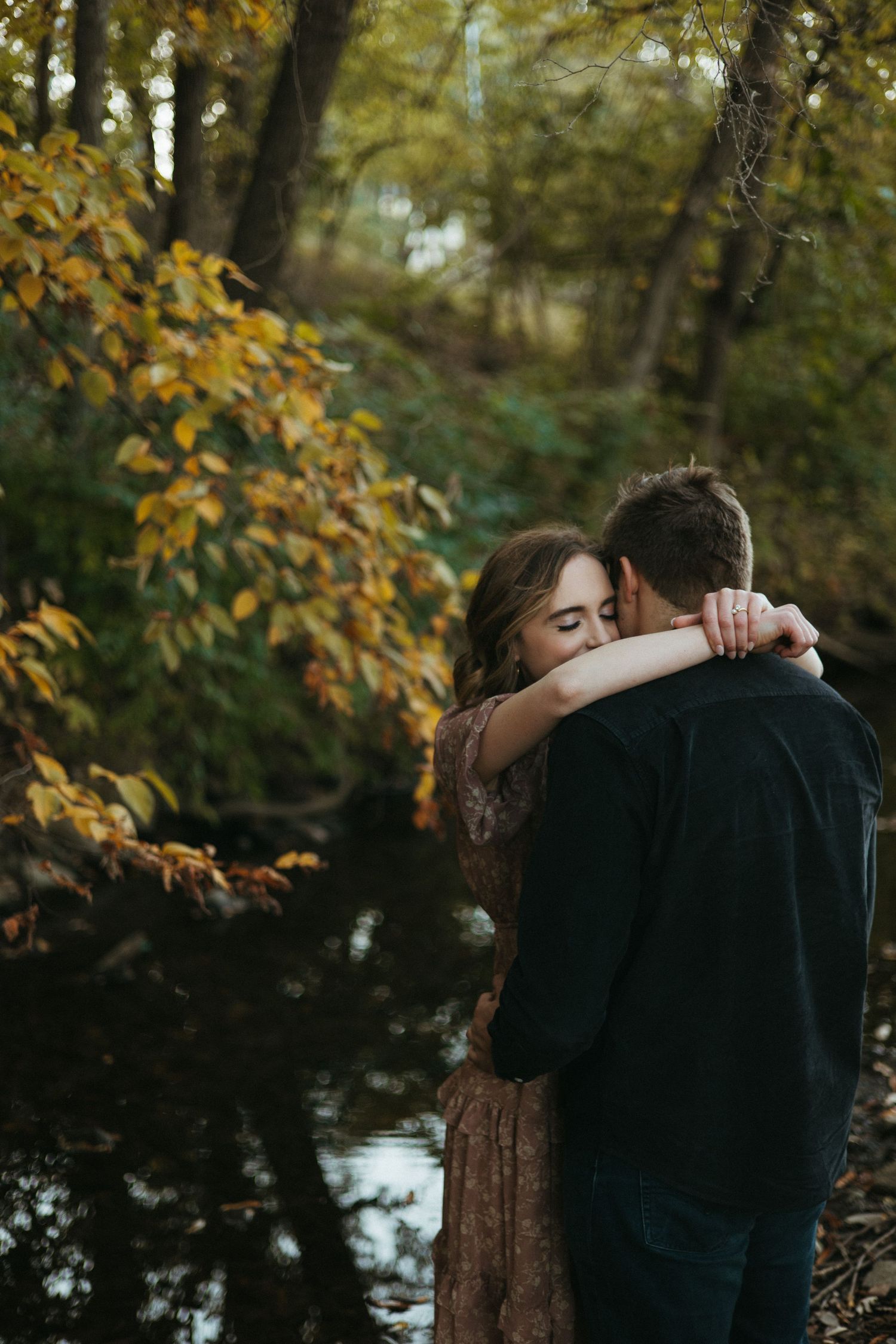 A couple embraces intimately in a wooded area with autumn foliage and a lake in the background.