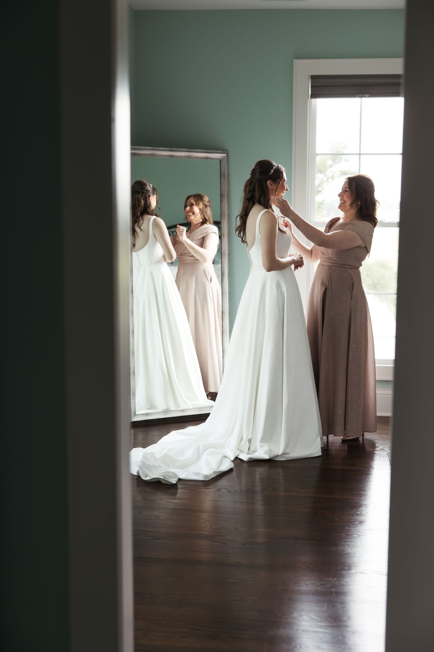 Bride in a white satin gown being buttoned up by a helper in front of a large mirror in a teal-walled dressing room.