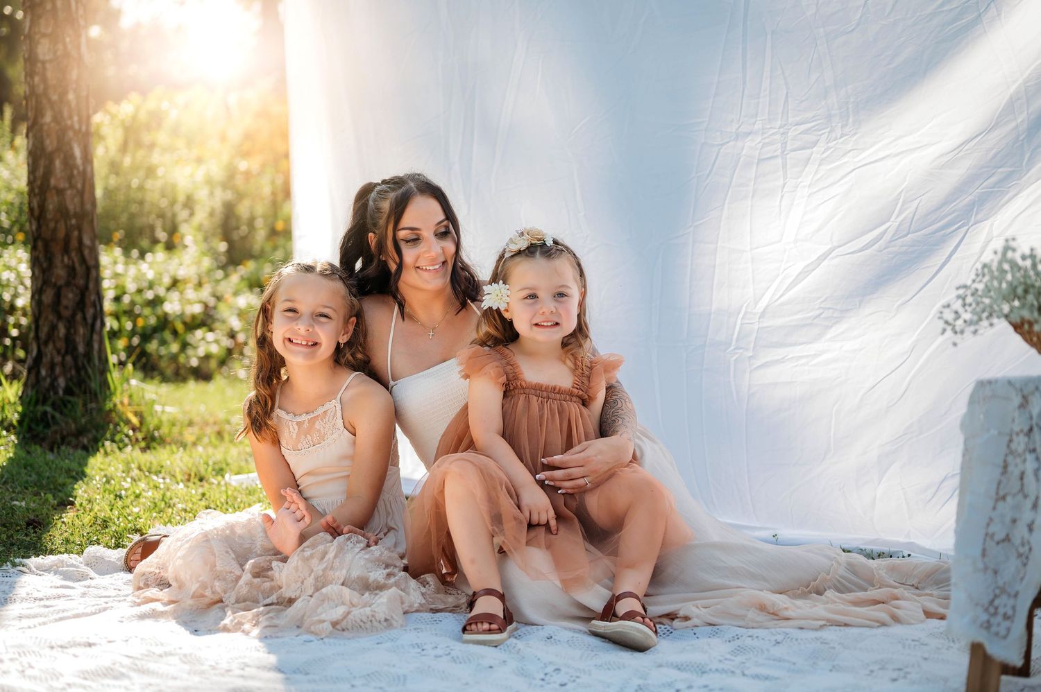 A woman sits outdoors with two young girls in matching neutral dresses against a white backdrop in soft sunlight.