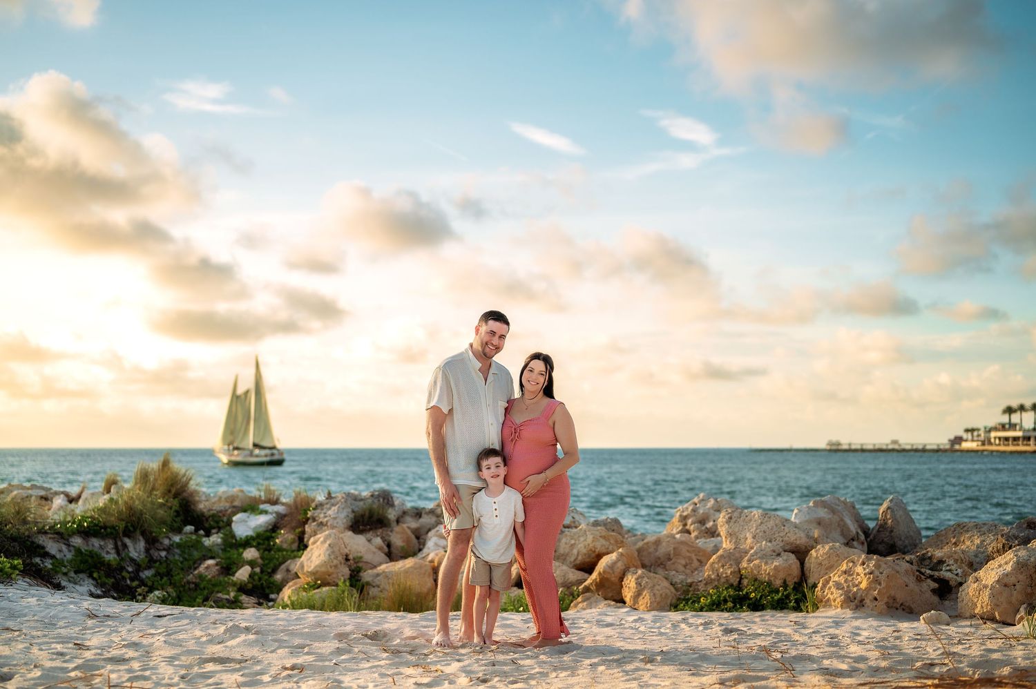 Family of three posing on a sandy beach at sunset with a sailboat and turquoise ocean in the background.