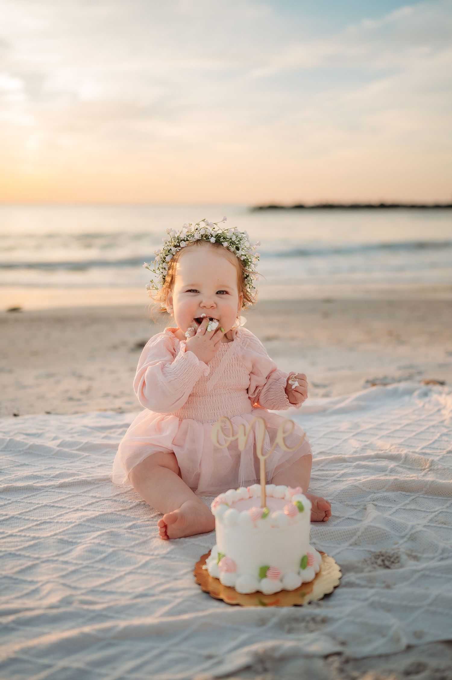Baby in pink dress with flower crown enjoying first birthday cake on beach at sunset.