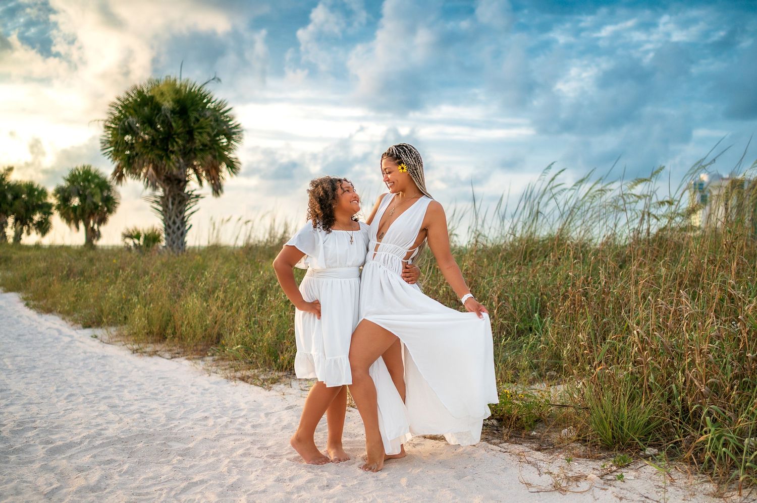 Mother and daughter in matching white dresses pose on a sandy beach path with palm trees and sea oats at sunset.