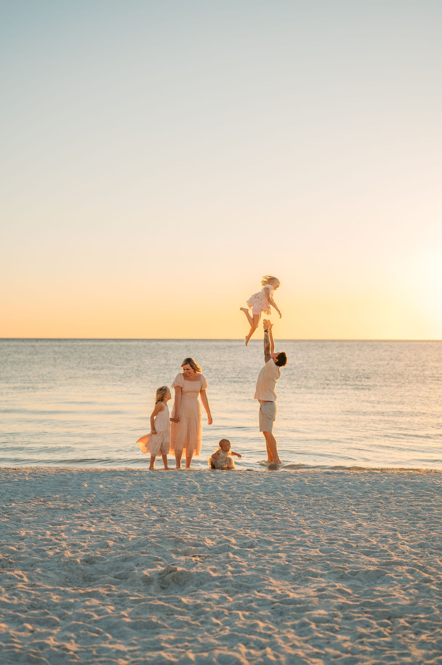 Family enjoying sunset on beach, father tossing child in air, mother and kids in matching cream outfits on white sand.