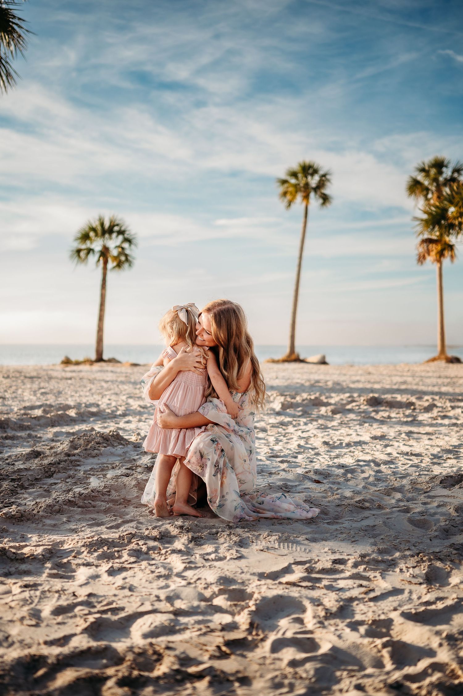Mom and daughter share an intimate moment sitting together on a sandy beach with palm trees and blue sky in the background.