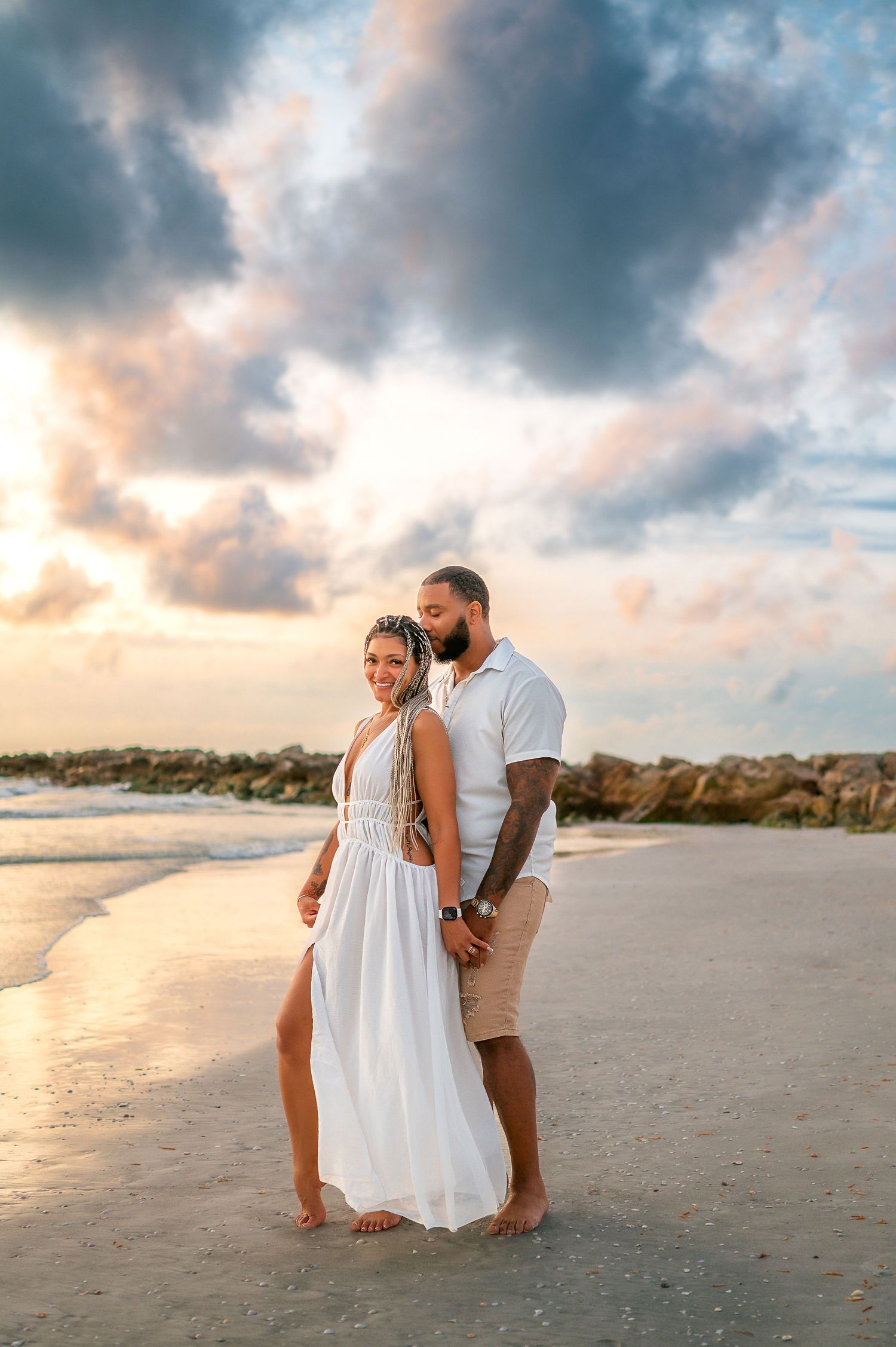 Couple embracing on a sandy beach at sunset, woman in white dress, man in white shirt with tattoos, dramatic cloudy sky.