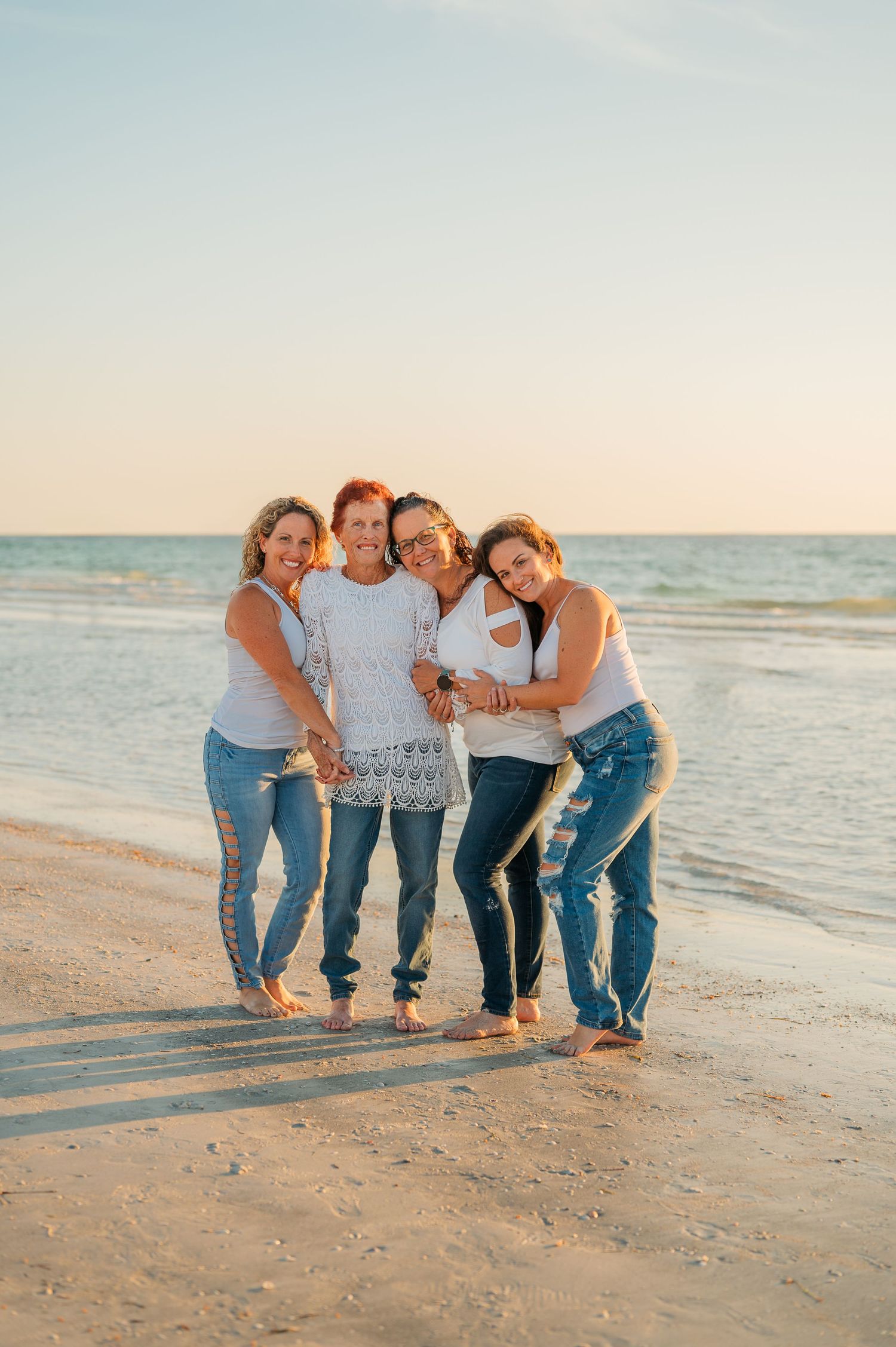 Four women in white tops and jeans smile together on a sandy beach at golden hour with ocean waves behind them.