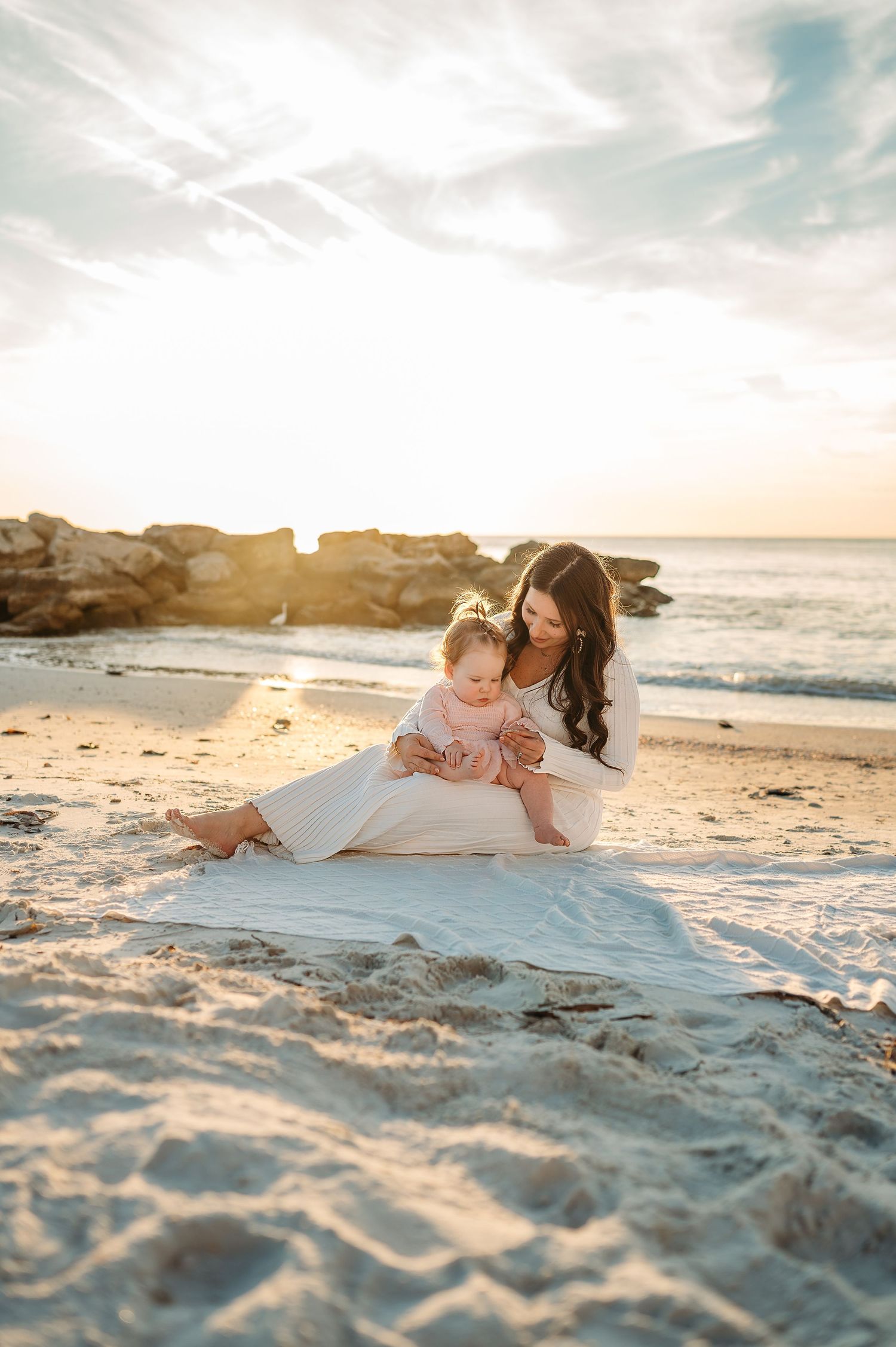 Mother and child sharing tender moment on sandy beach at sunset near rocky shore.