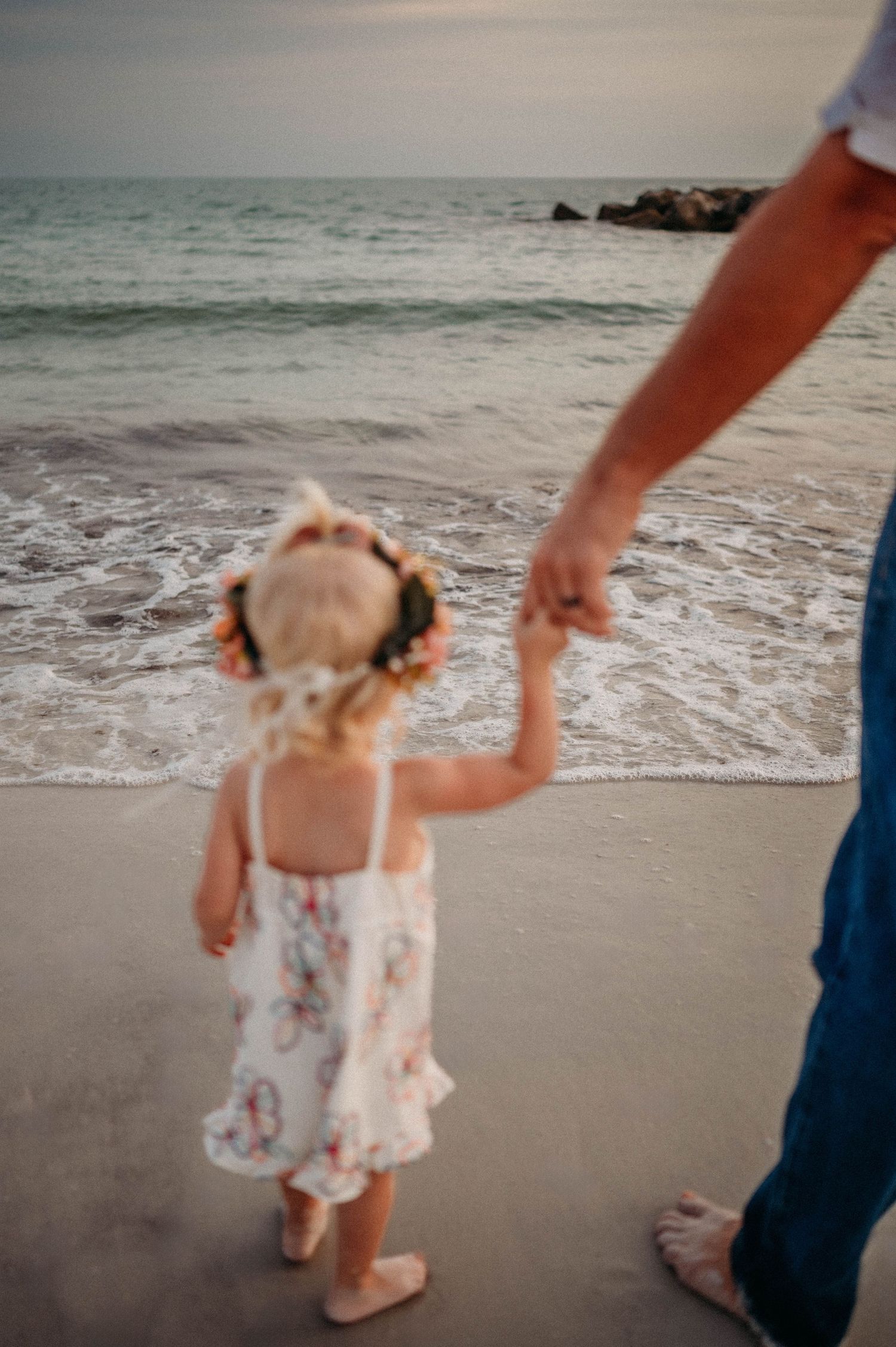 Child in floral dress walking on beach at sunset holding hands with father.
