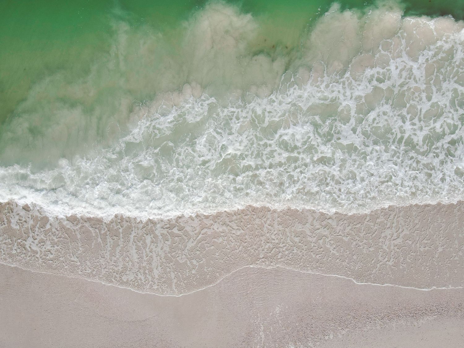 Aerial view of ocean waves crashing onto a sandy beach with white sea foam patterns.