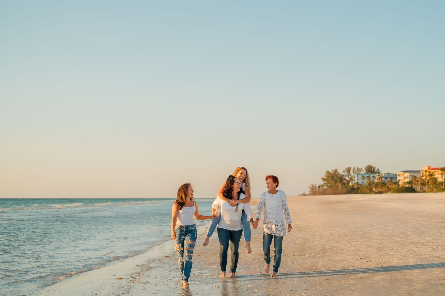 Family of four walking barefoot on a sunny beach at sunset, smiling and holding hands near the shoreline.