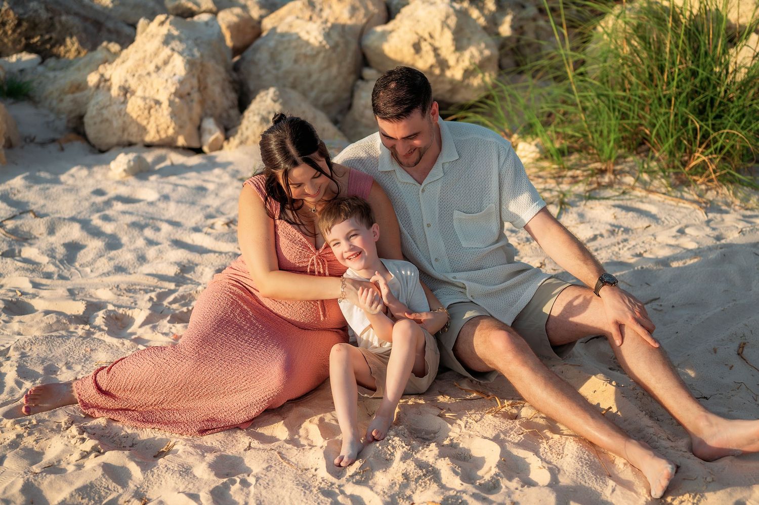 Happy family of three relaxing on sandy beach at golden hour, parents cuddling young child near rocky shore.