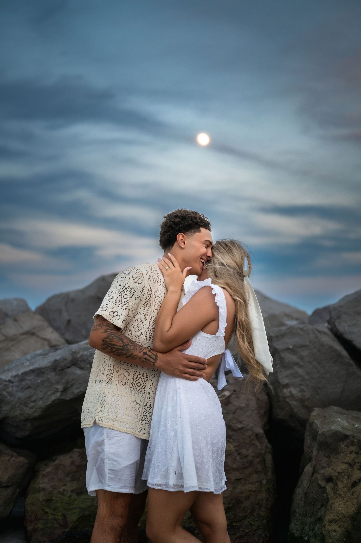 Couple in white clothing embracing against rocky coastline at dusk.