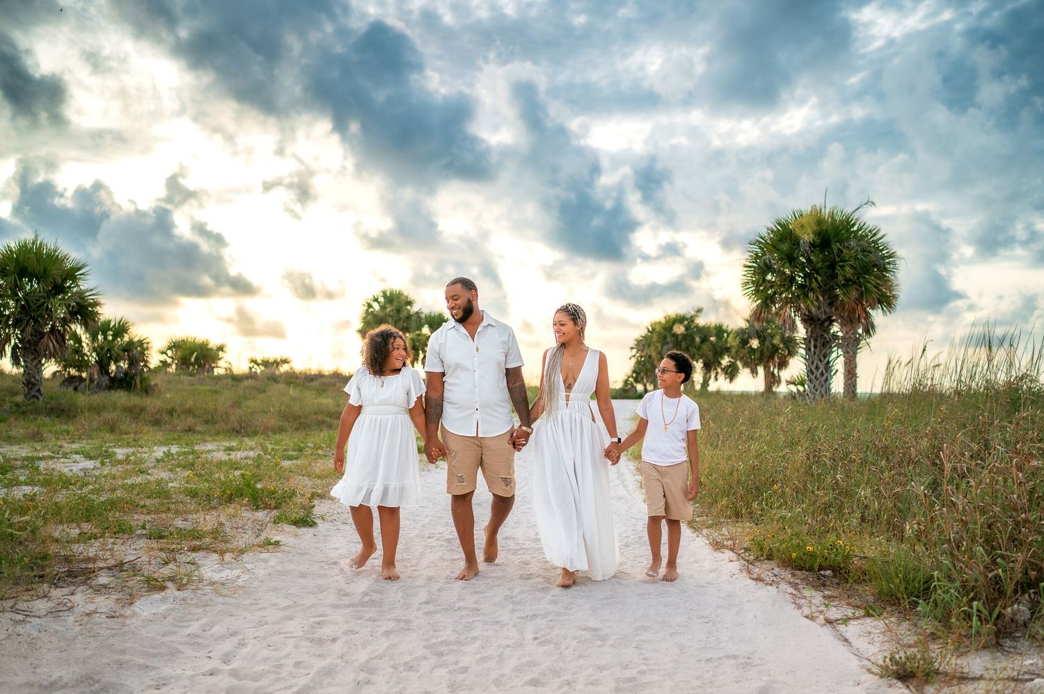 Family in white outfits walking on sandy beach path at sunset with palm trees and dramatic cloudy sky.