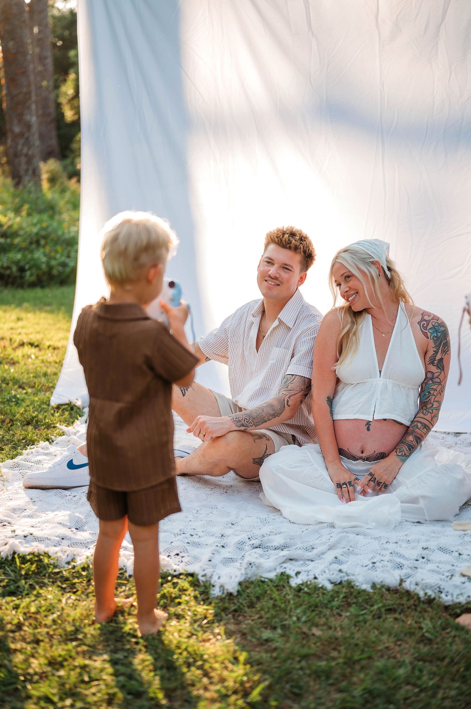 A young boy photographs a smiling couple sitting on a blanket outdoors, the woman visibly pregnant with tattoos.