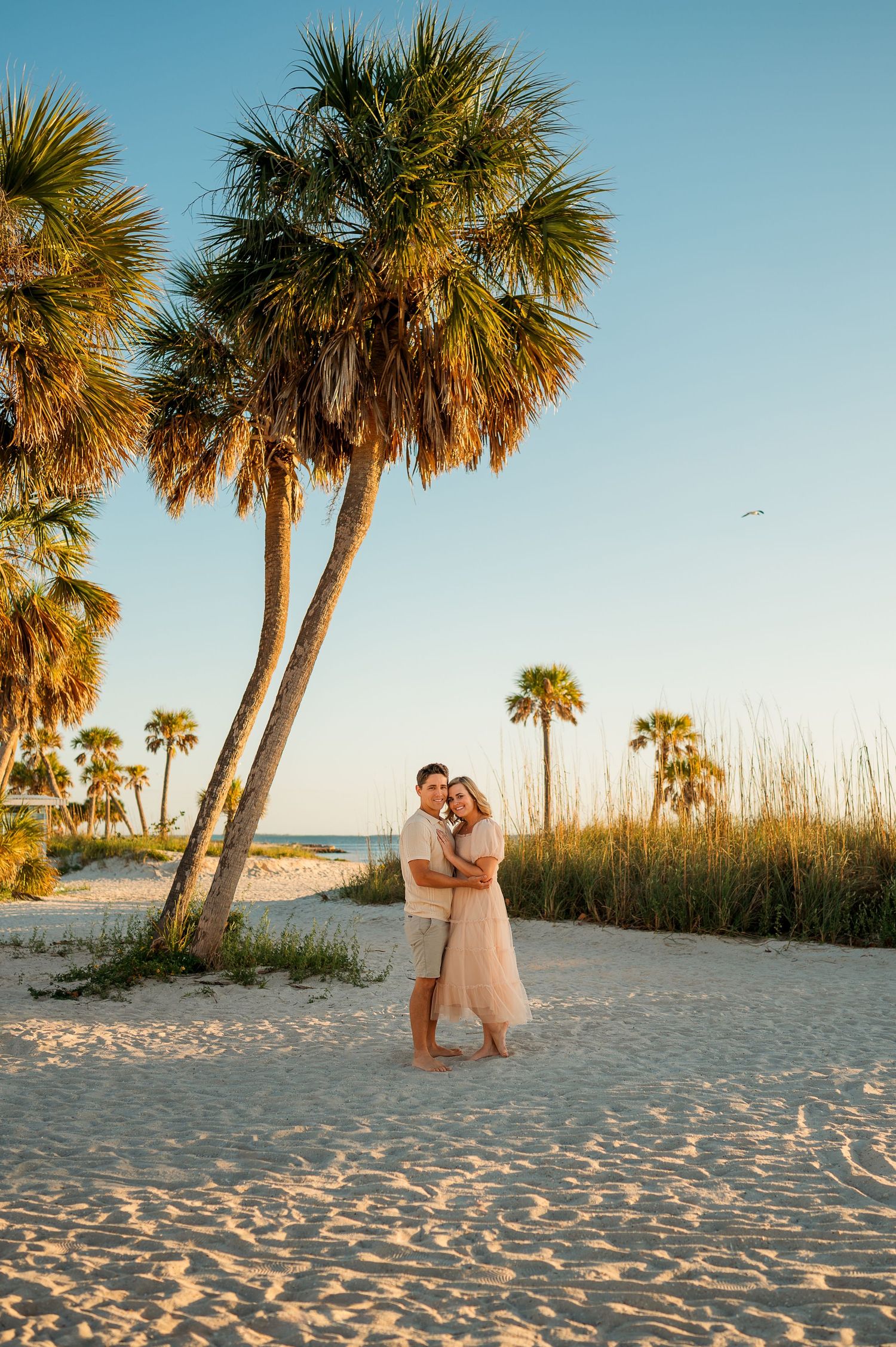 Couple embracing on white sand beach at golden hour with tall palm trees and blue sky in background.