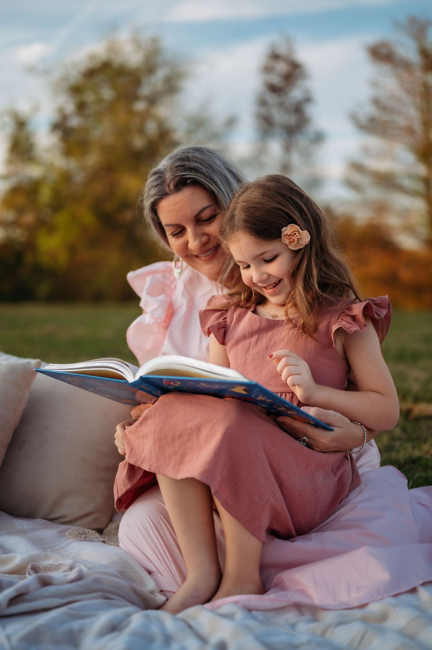 Grandmother and grandchild in pink dresses reading together on a blanket in a park during golden hour.