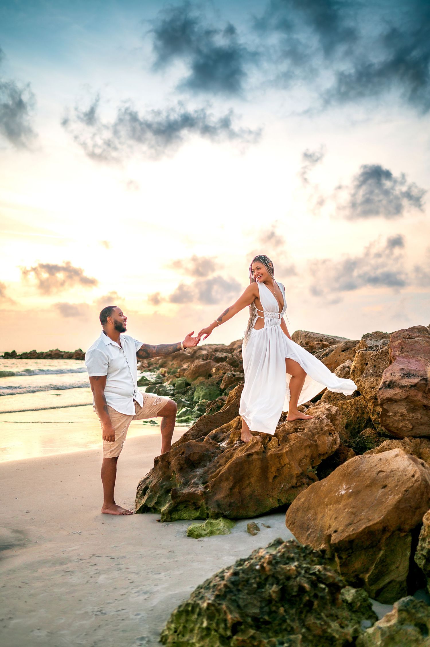 Couple in white outfits holding hands on rocky beach at sunset, waves and dramatic sky in background.