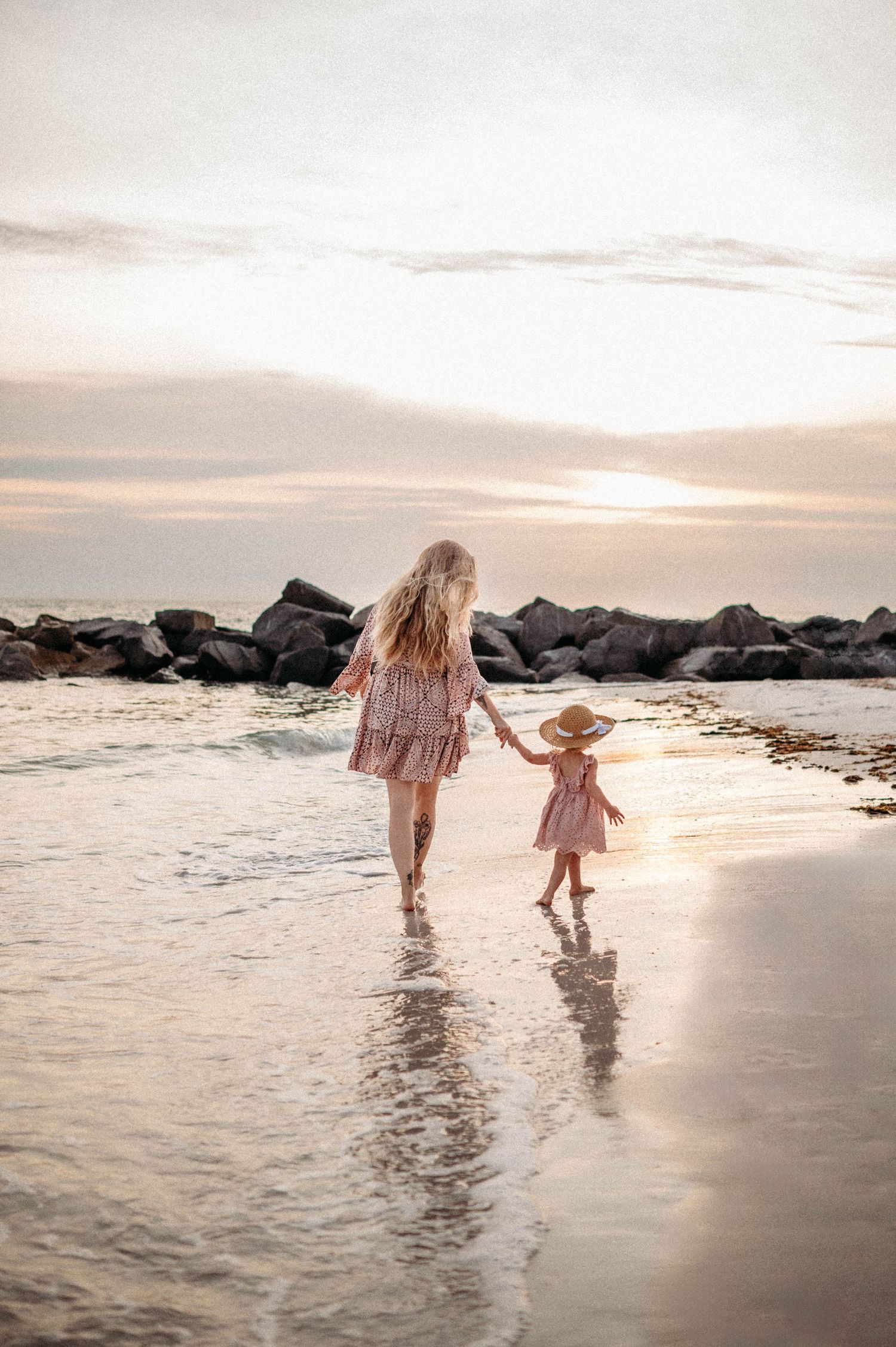 Mother and child walk hand in hand through shallow ocean waves at sunset.