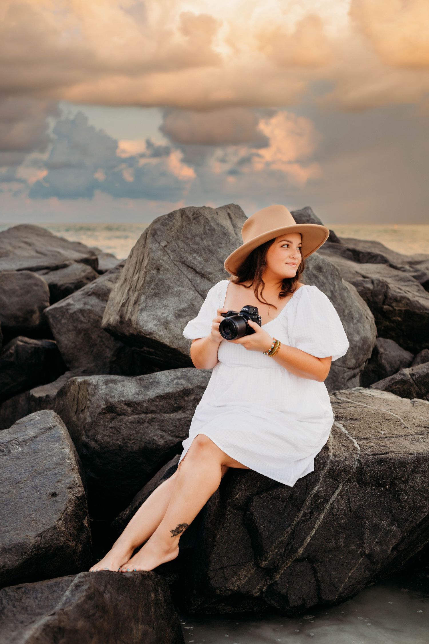 A figure in a white dress and sun hat reclines on dark coastal rocks during golden hour sunset.