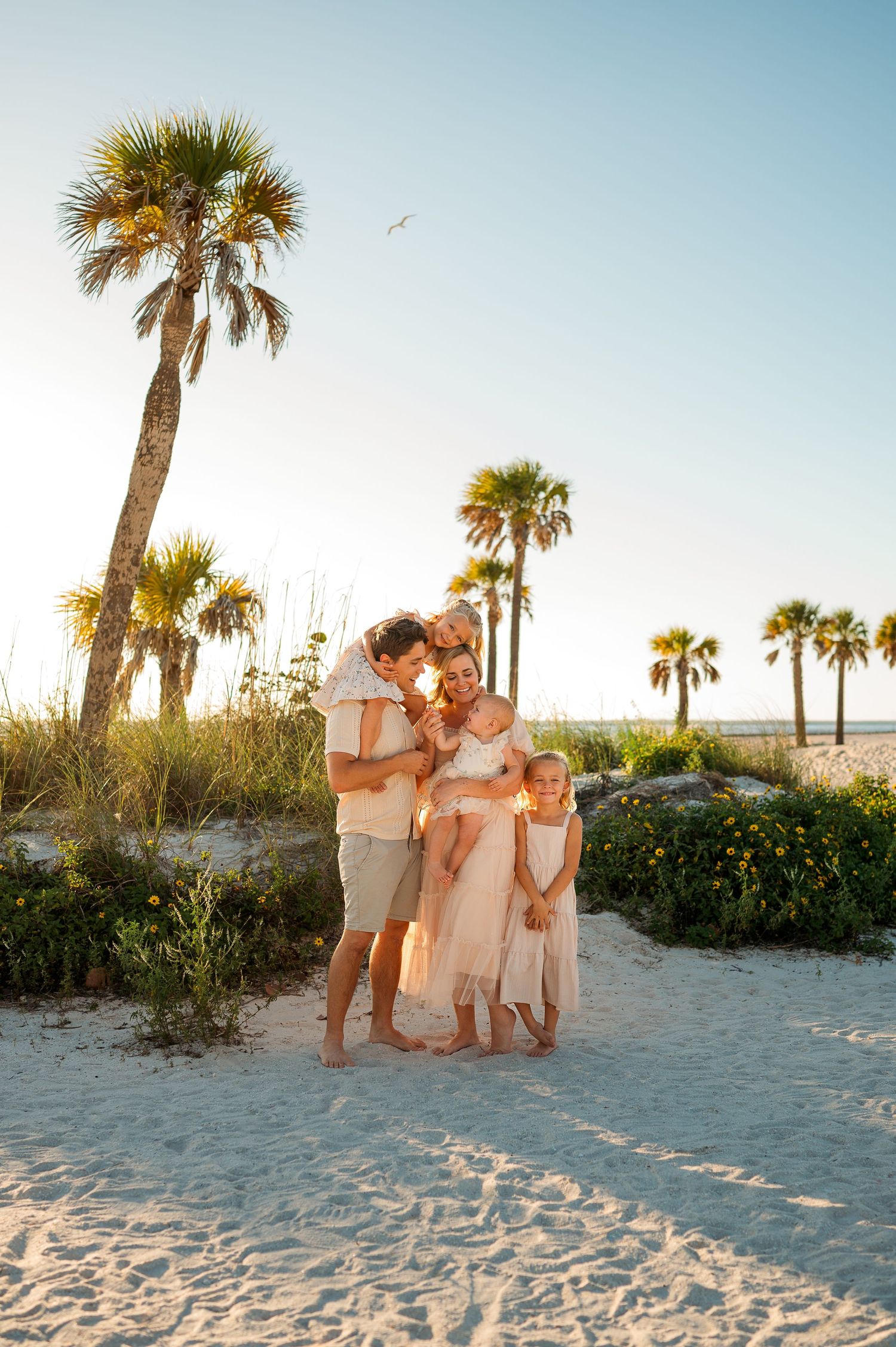 Family of four poses barefoot on white sand beach at sunset with palm trees in warm golden light.