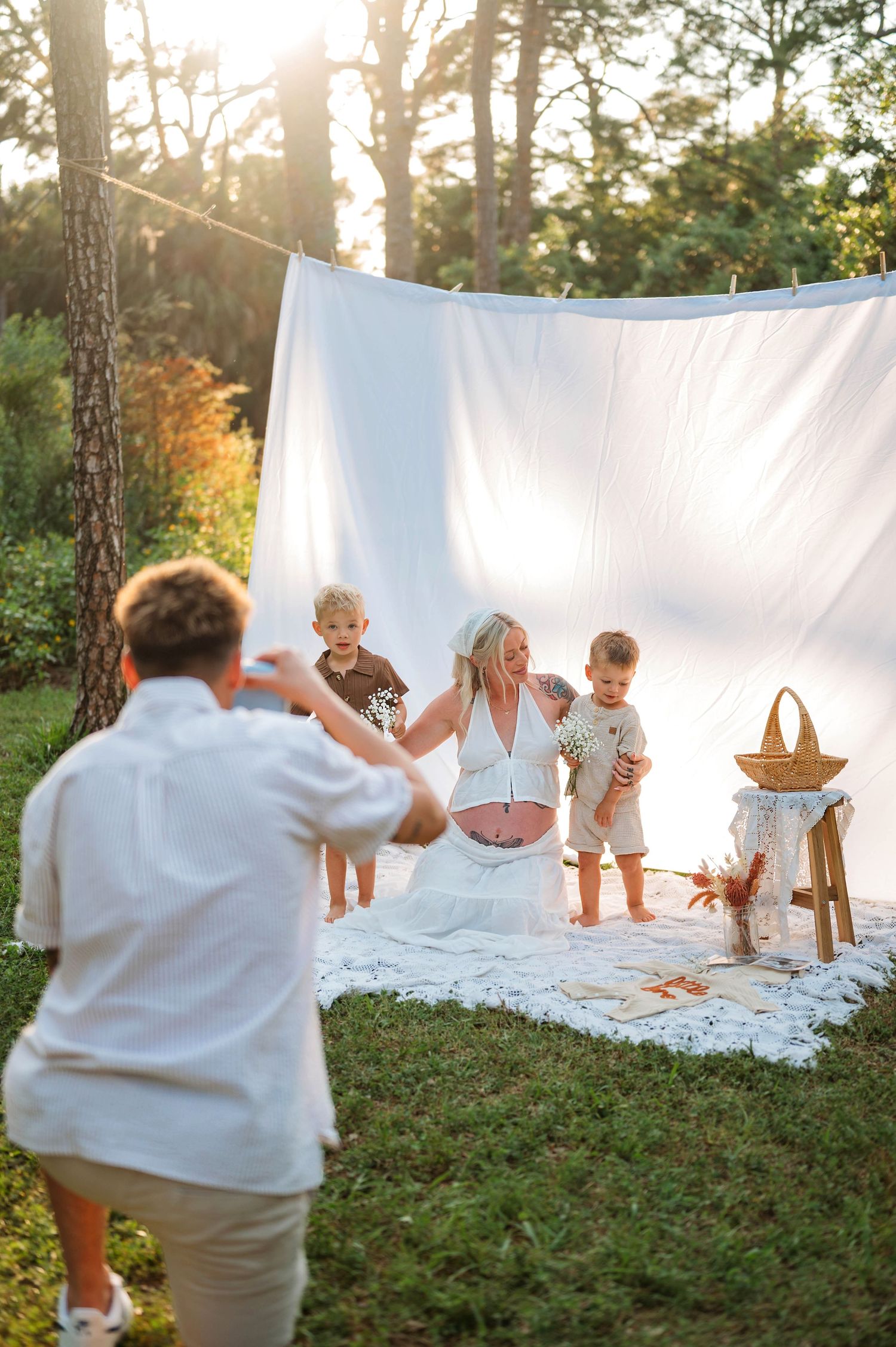 A man photographs a pregnant woman with two toddlers against a white backdrop outdoors at golden hour.