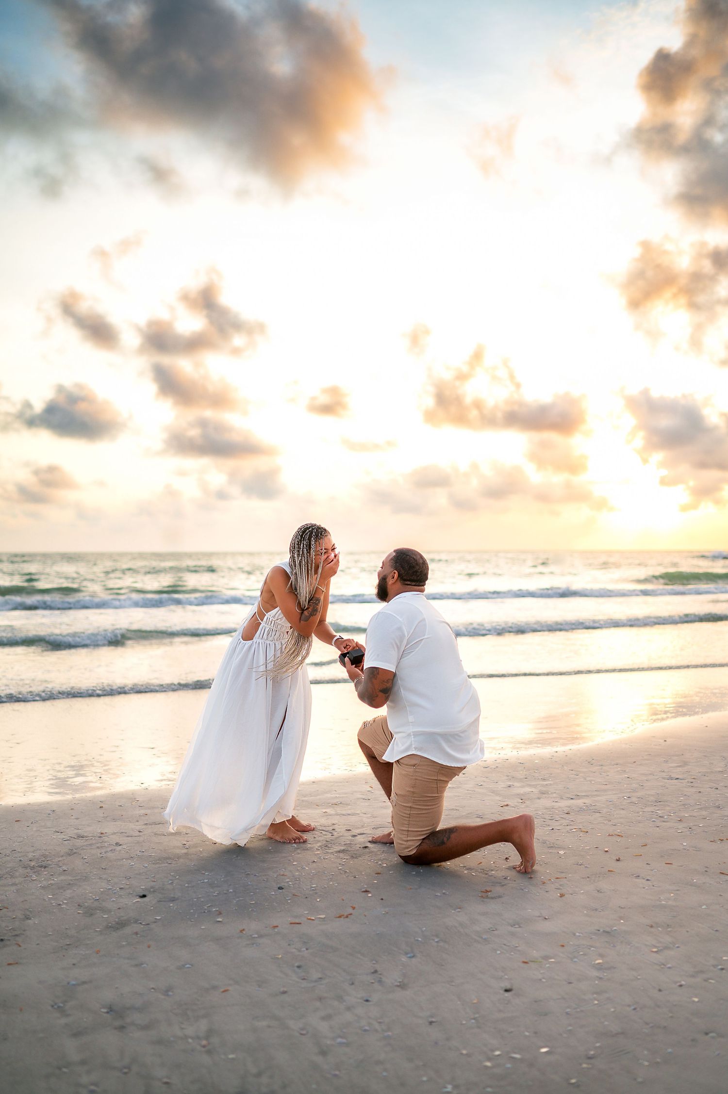 Man proposes on bended knee to woman in white dress on a beach at sunset with dramatic clouds.