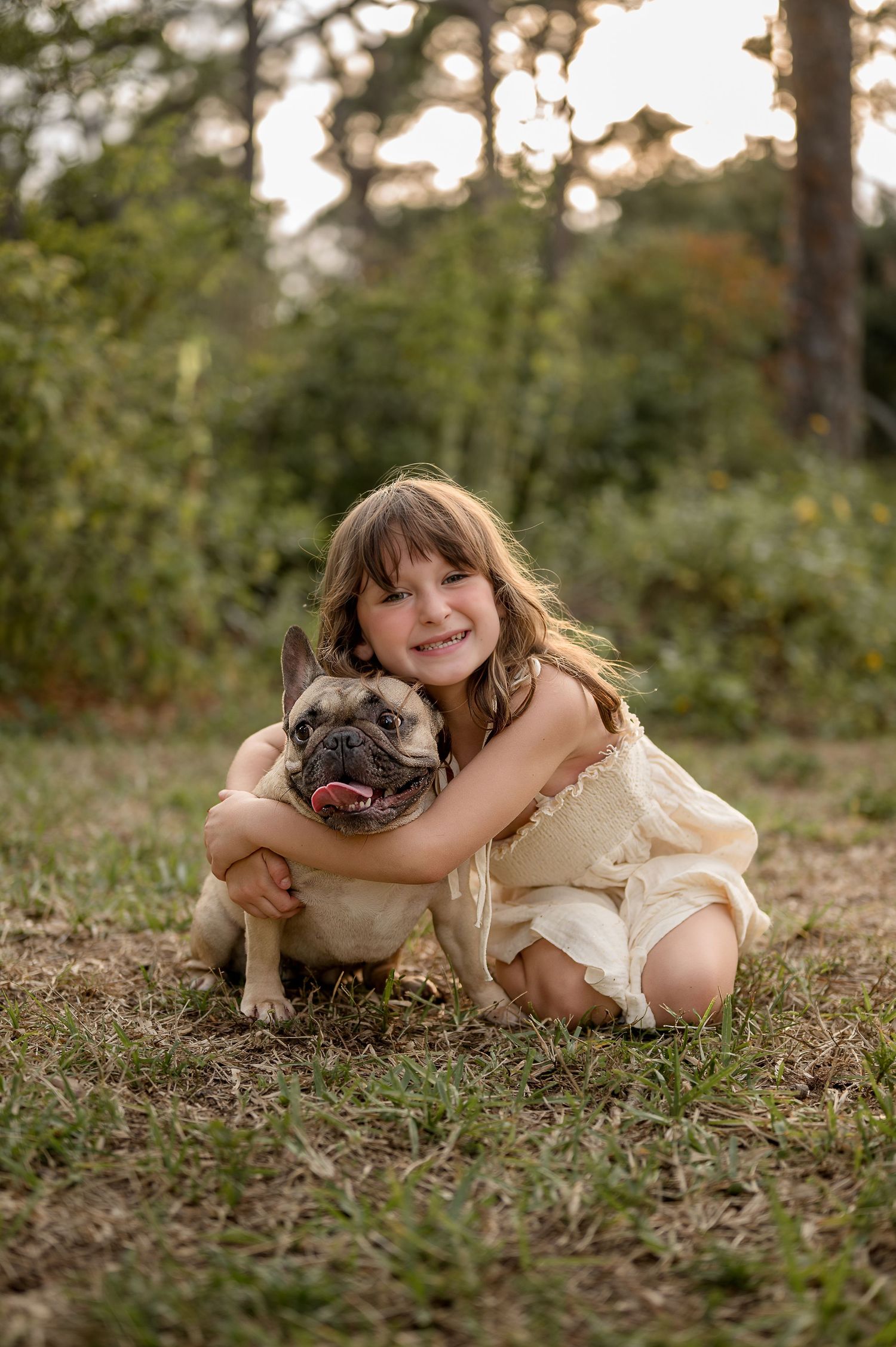 A smiling girl in a cream dress hugs a French Bulldog outdoors in a sunlit green park setting.