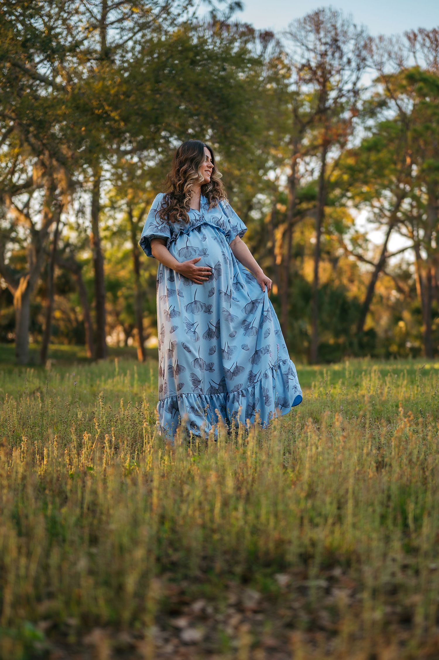 Maternity photoshoot, woman in flowing blue floral dress twirls gracefully in a sunlit meadow during sunset.
