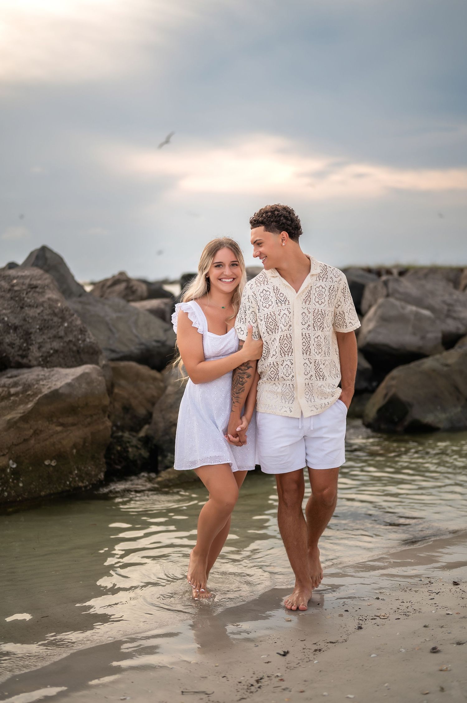 A newly engaged couple in white outfits shares joyful moments along a rocky beach at sunset.