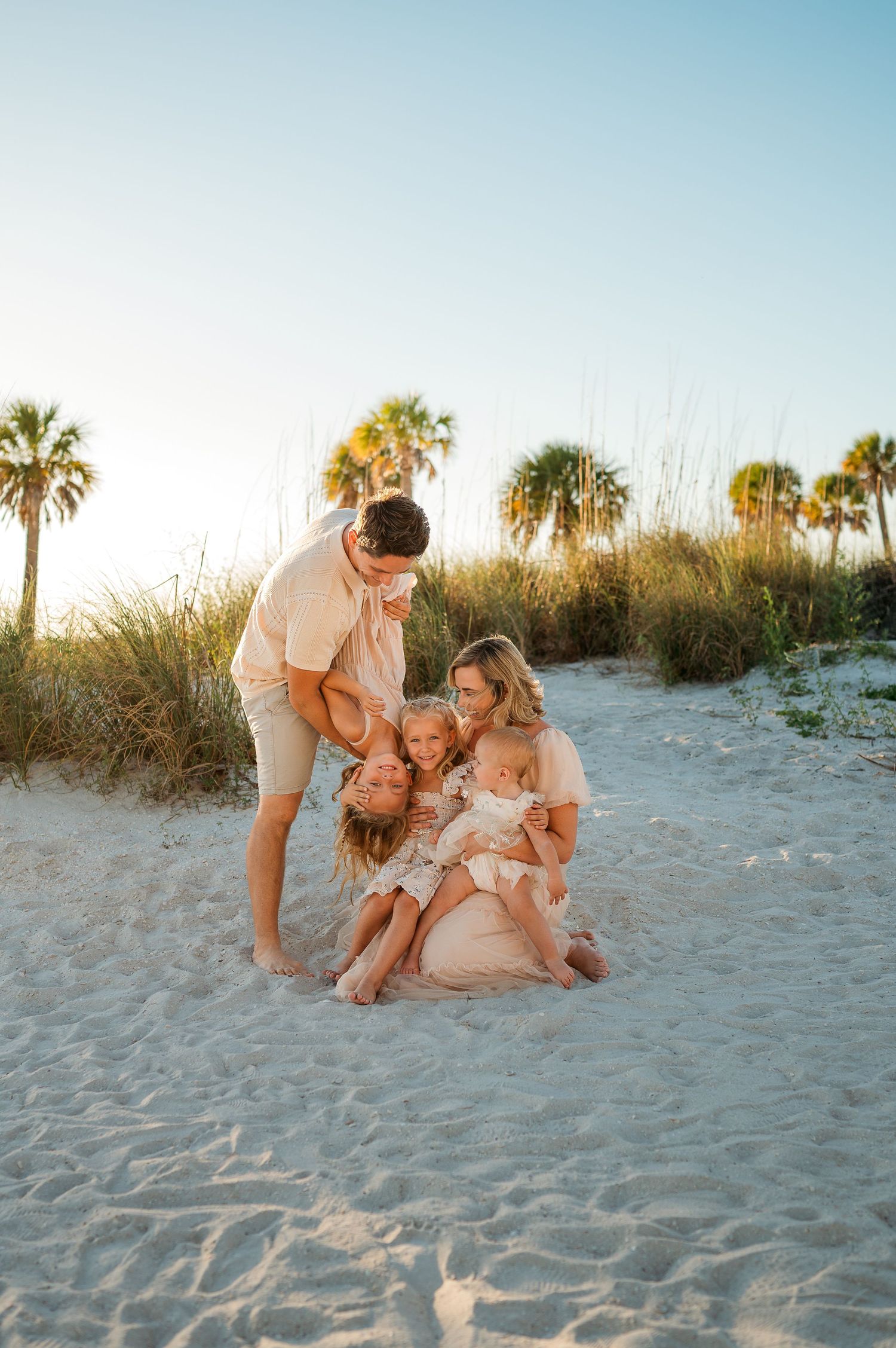 Family of five sharing a joyful moment on a white sand beach at golden hour with palm trees backdrop.