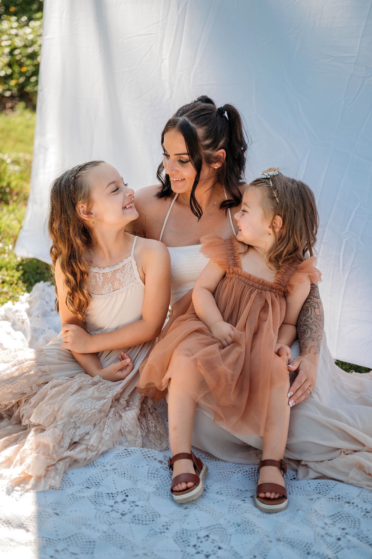 A mother and her two young daughters in elegant dresses share a joyful moment sitting together outdoors.