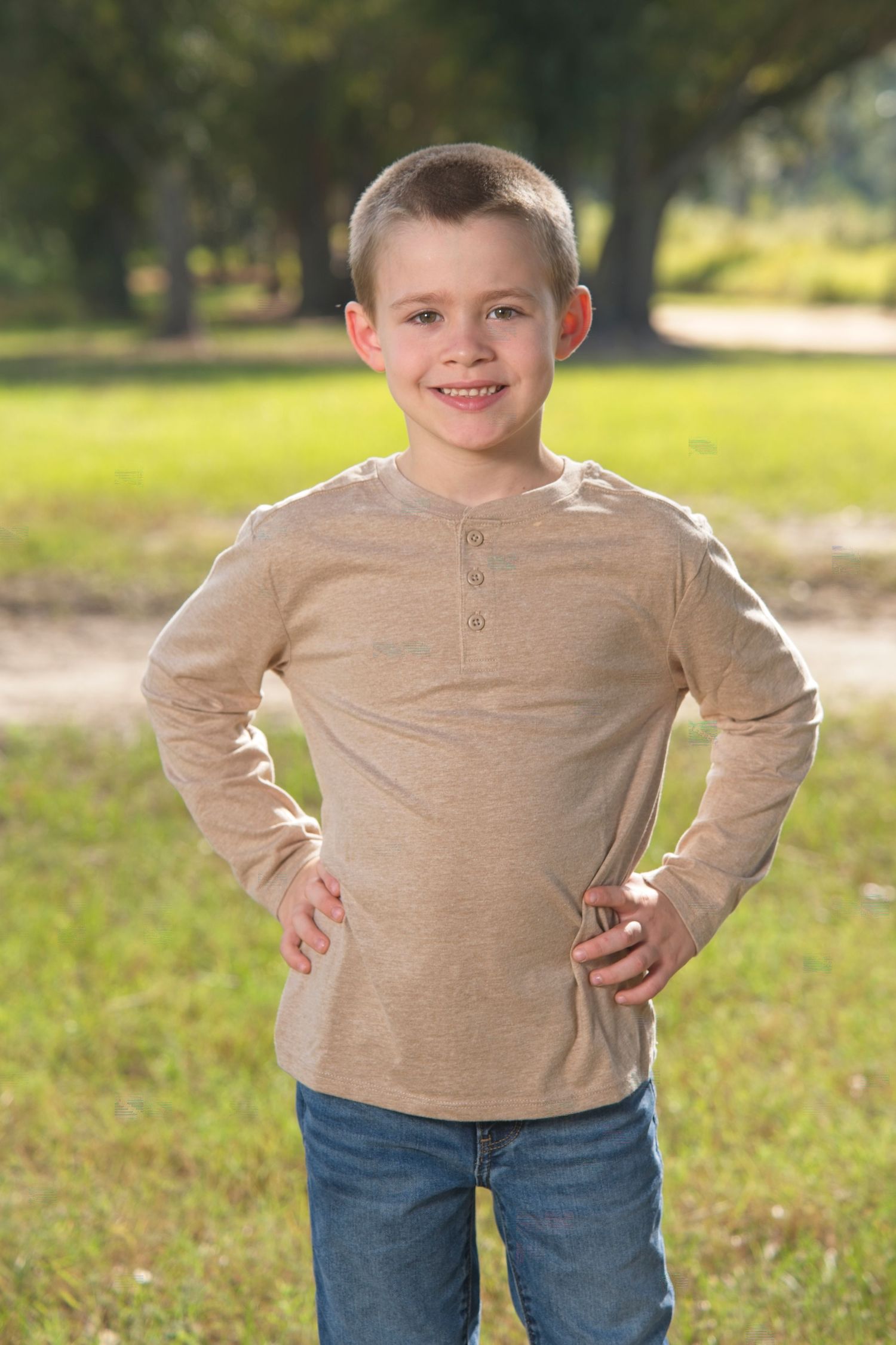 A child wearing a beige sweater and blue jeans stands outdoors in a grassy field on a sunny day.
