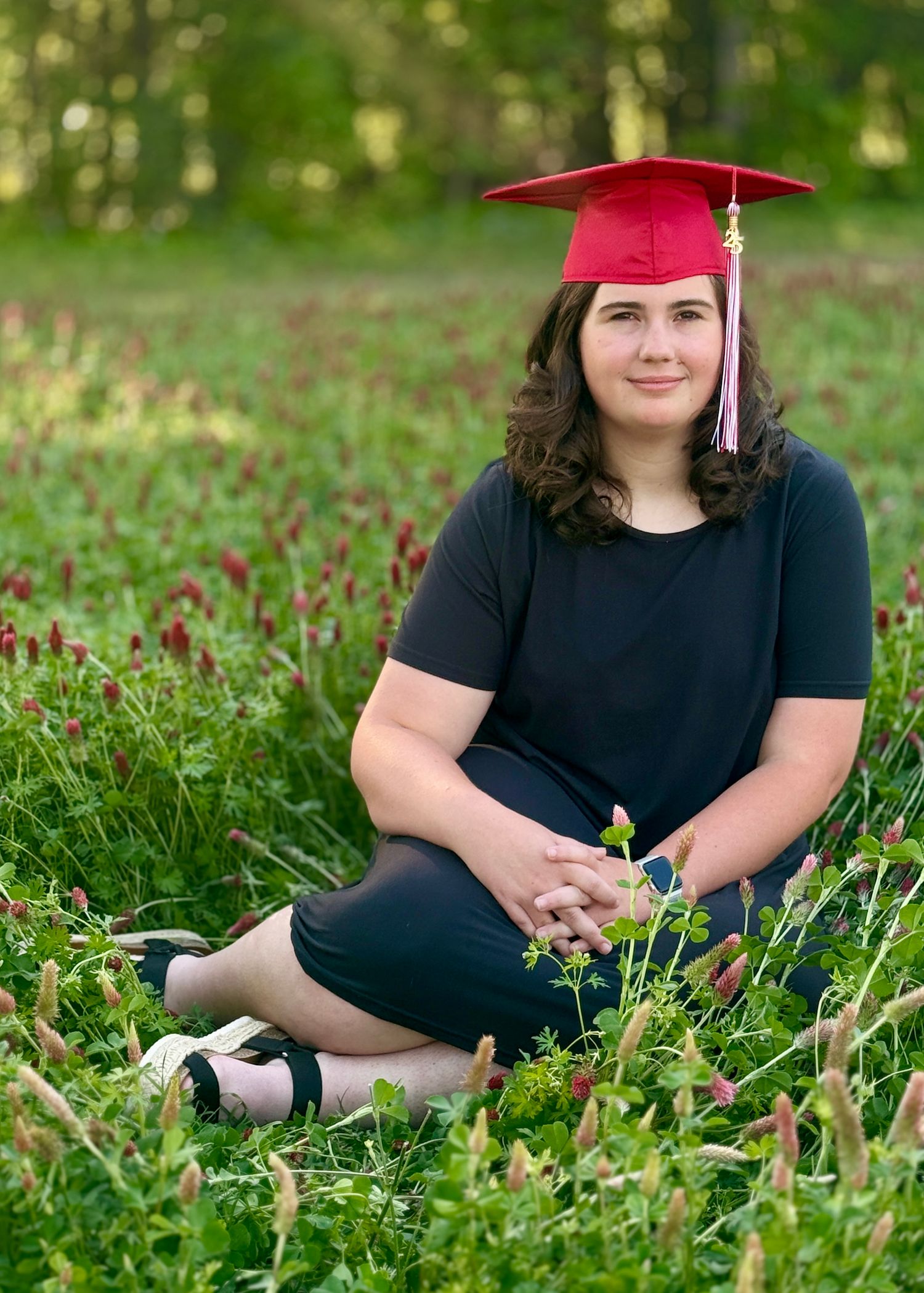 Graduate in red cap sitting in a field of wildflowers wearing black dress during outdoor portrait session.