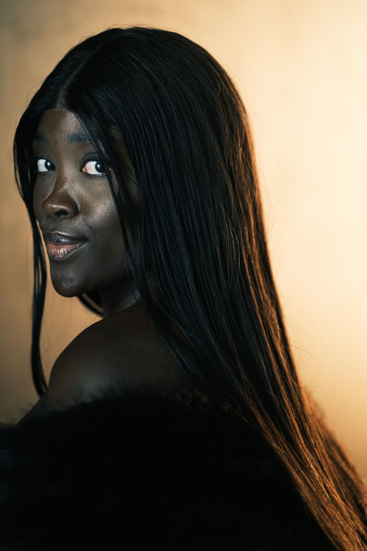 A Black woman with long straight hair poses dramatically against a warm golden studio background.