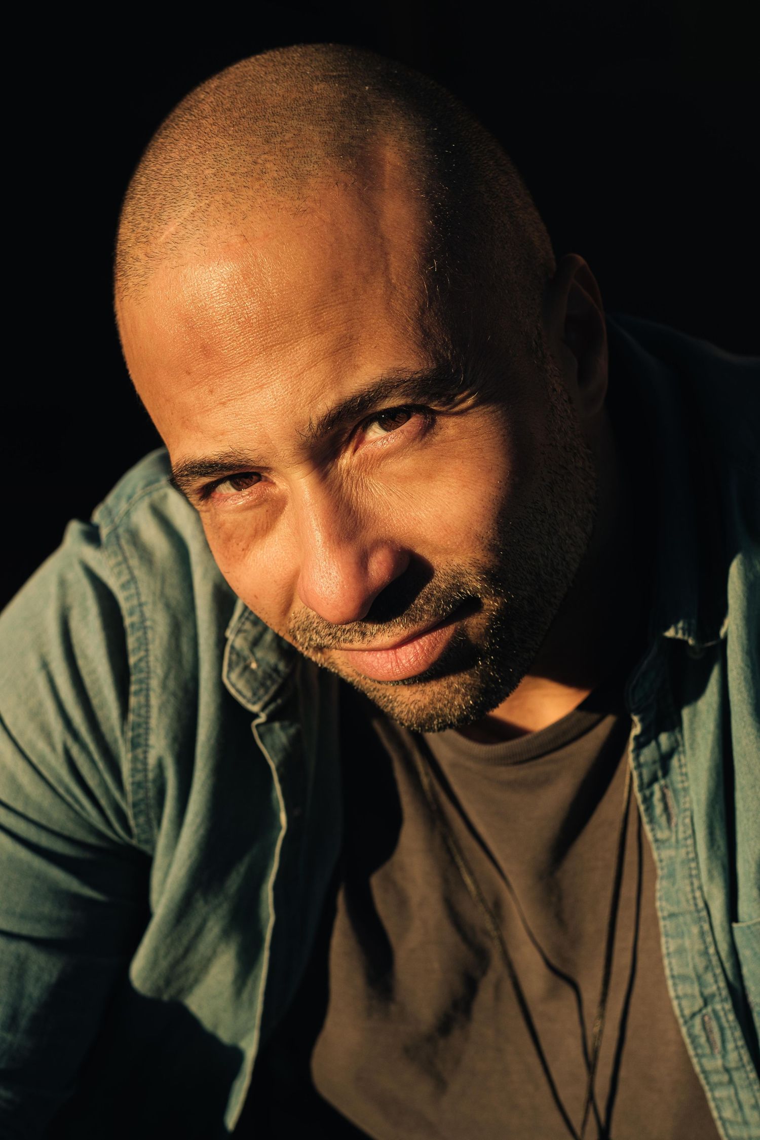 A bald man with a beard in a denim shirt poses in dramatic side lighting.