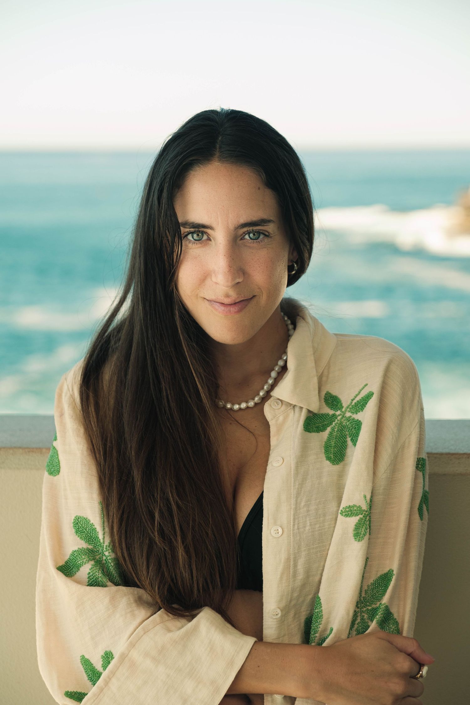 Young woman with long dark hair wearing a floral shirt and pearl necklace, posing on a balcony with ocean views.