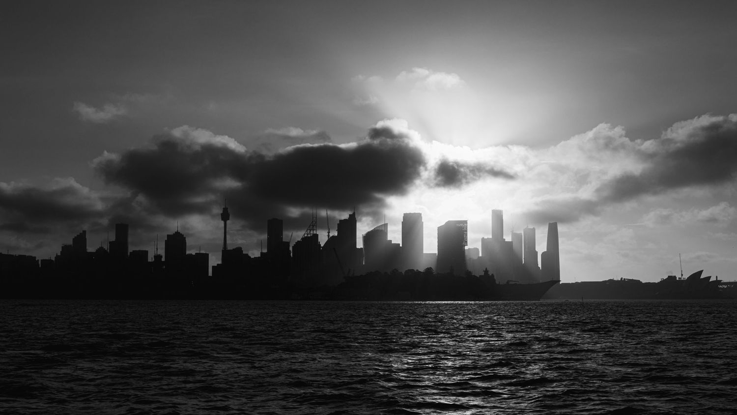 Dramatic black and white cityscape silhouette against a cloudy sky over water.
