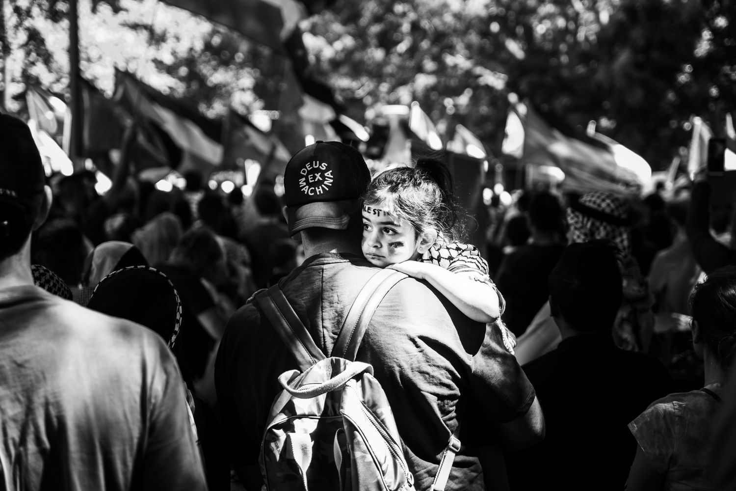 Person carrying child on shoulders in a crowd at an outdoor protest with flags.