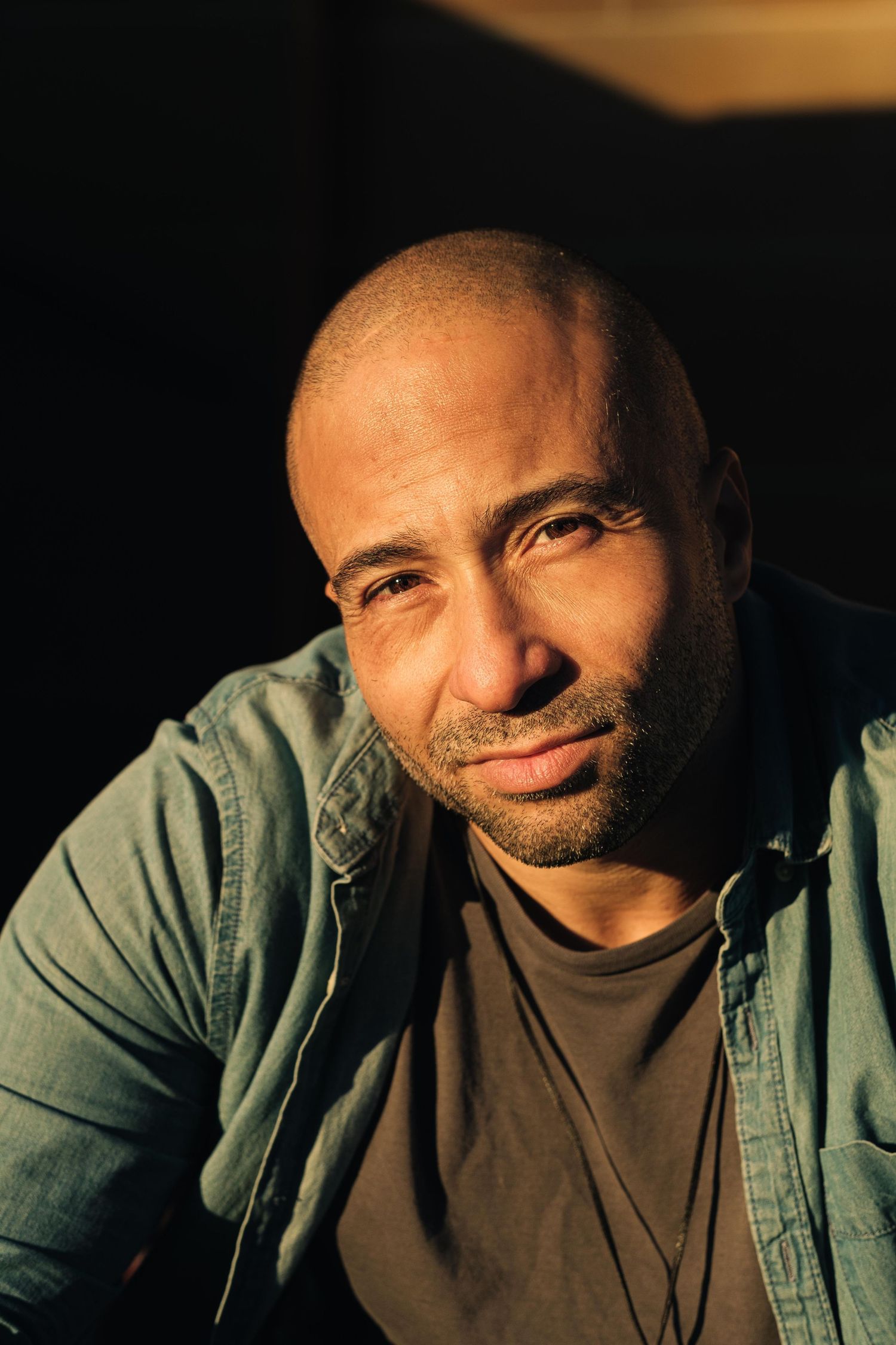 A bald bearded man in a denim shirt smiles warmly in dramatic golden side lighting.