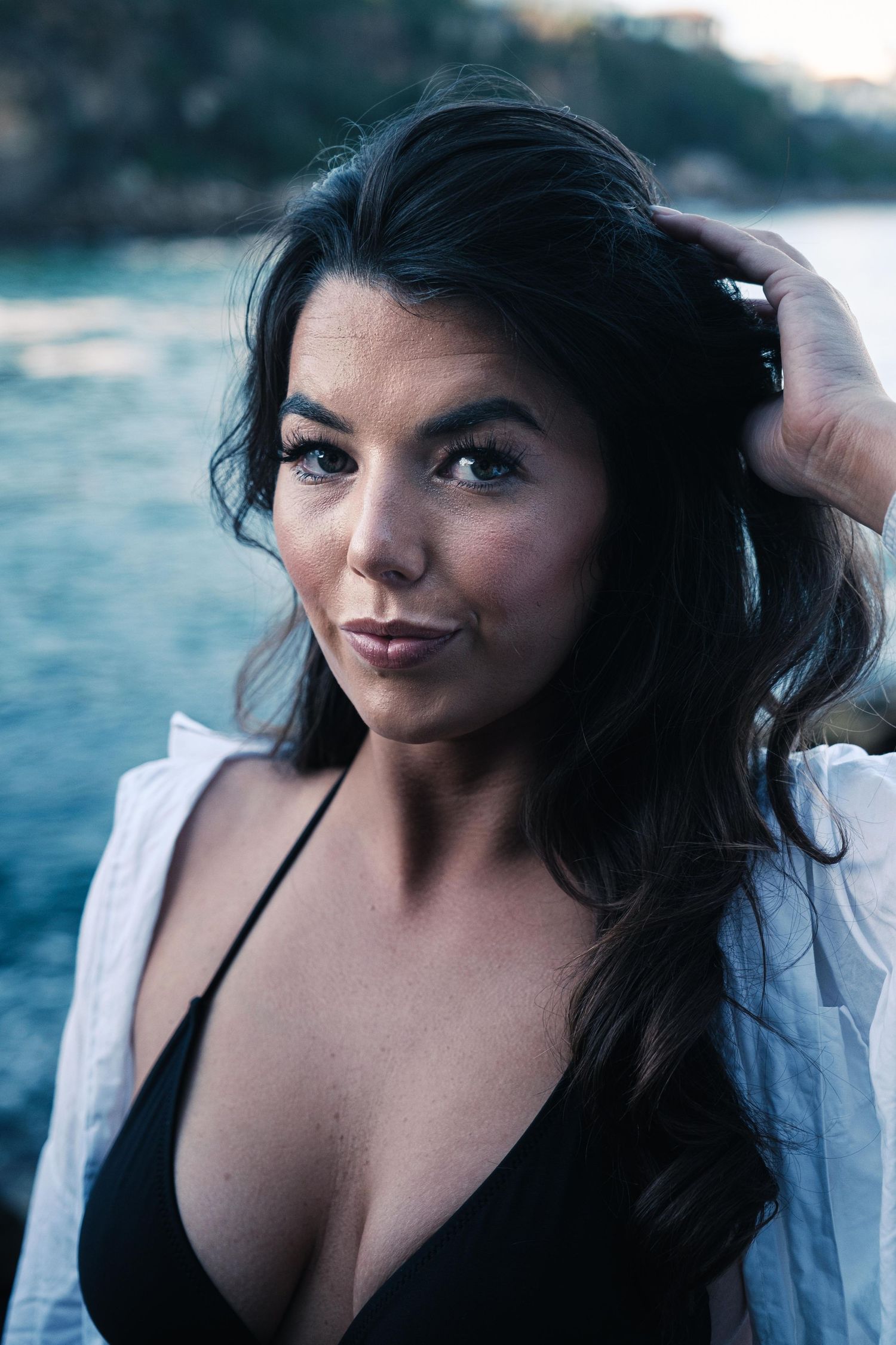 Dark-haired woman in a black bikini top and white shirt posing near the ocean at dusk.