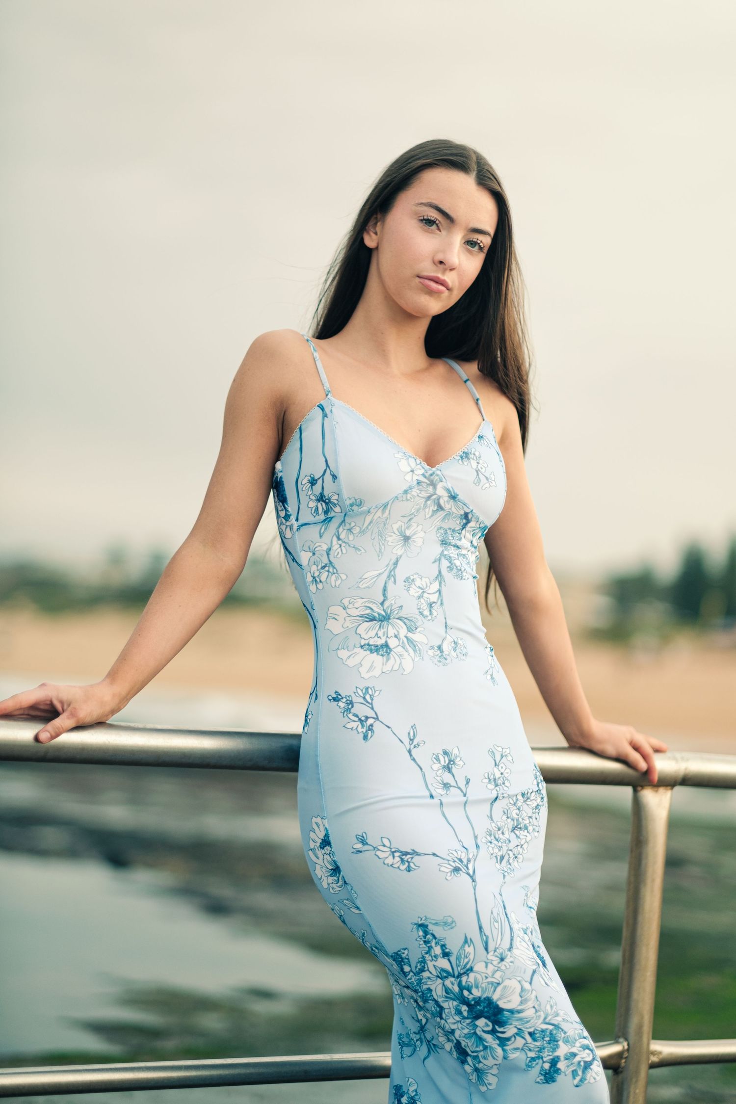 Stunning young woman in a fitted blue floral maxi dress leaning against a railing outdoors.