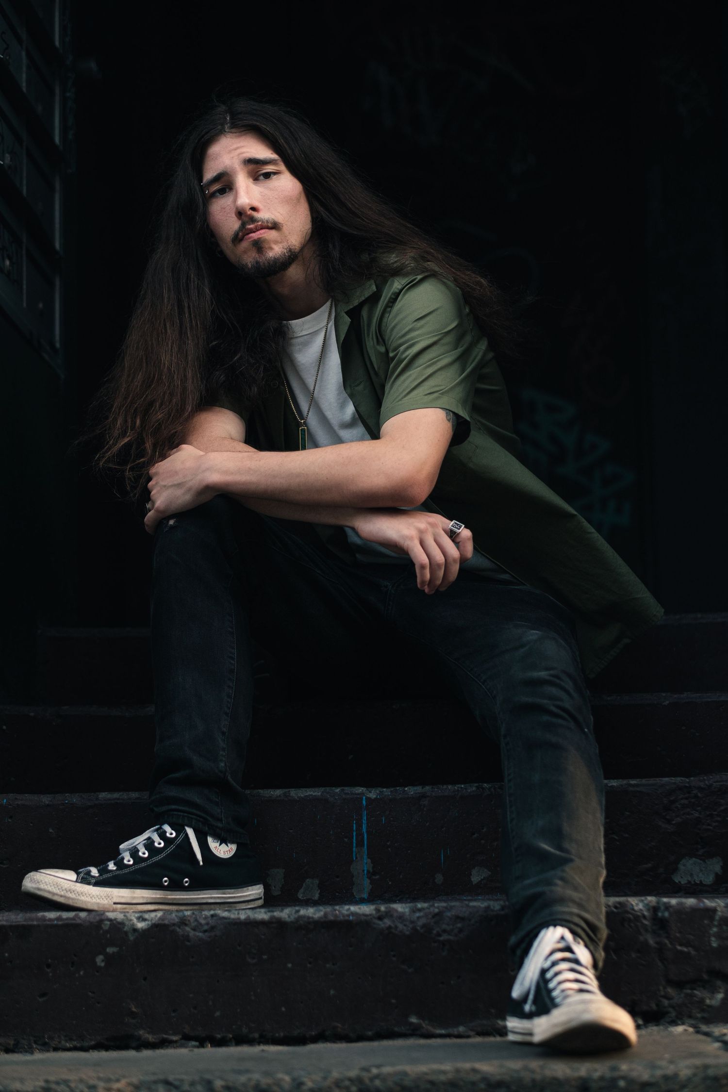 Young man with long dark hair sitting on steps in a green jacket and black jeans outdoors.