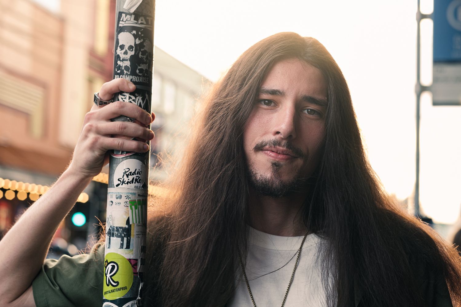 A young man with very long hair holds a sticker-covered pole on a city street.