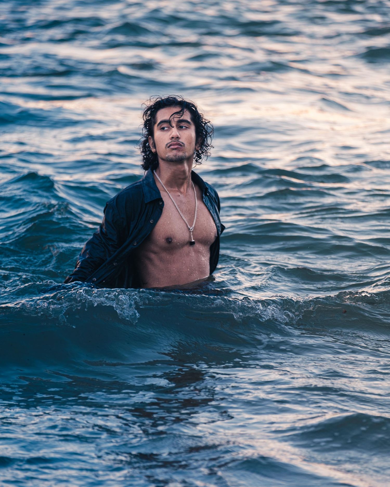 Young man with curly hair standing waist-deep in ocean waves wearing an open dark shirt.