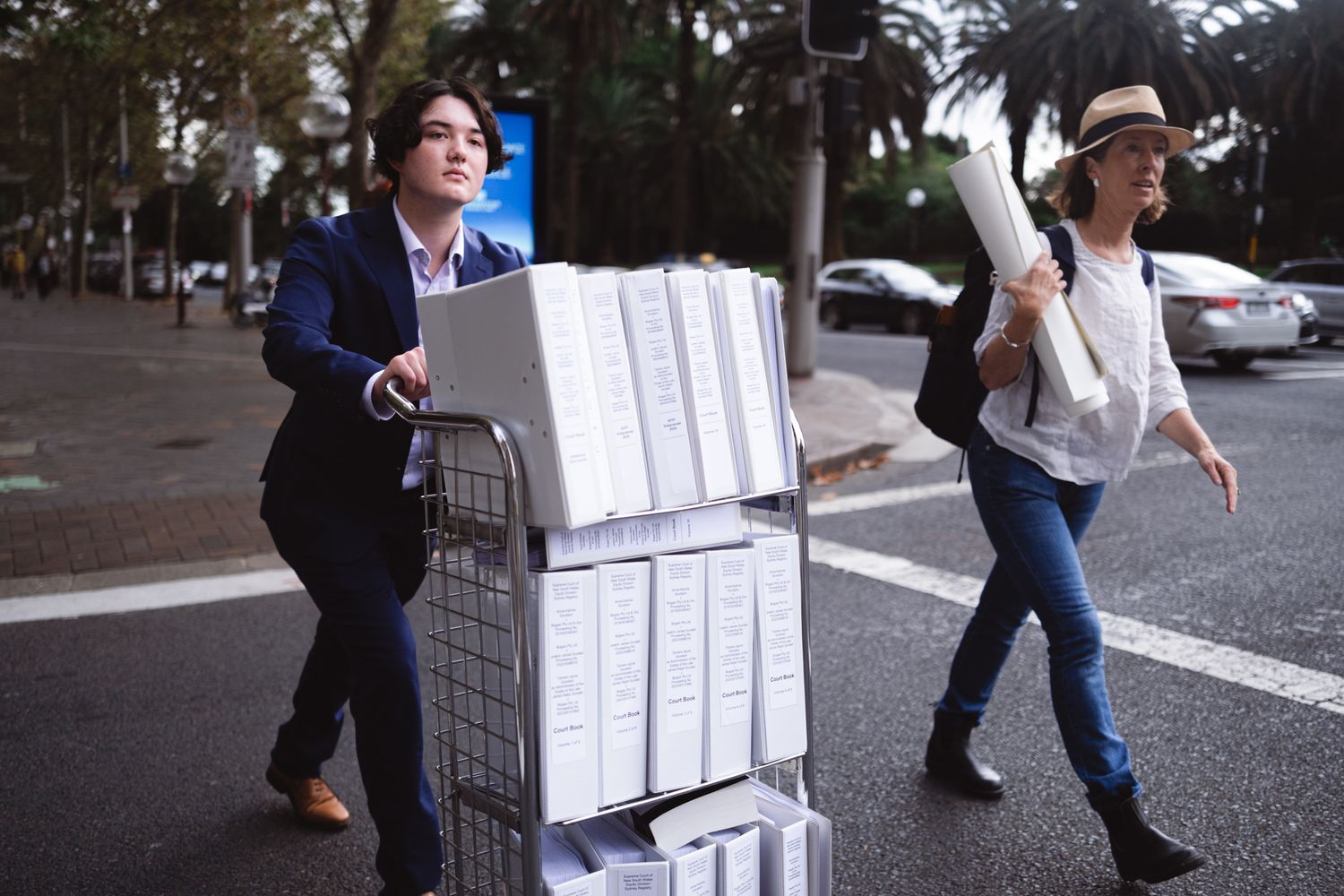 Two people crossing a road, one pushing a trolley stacked with white binders, other carrying plans.